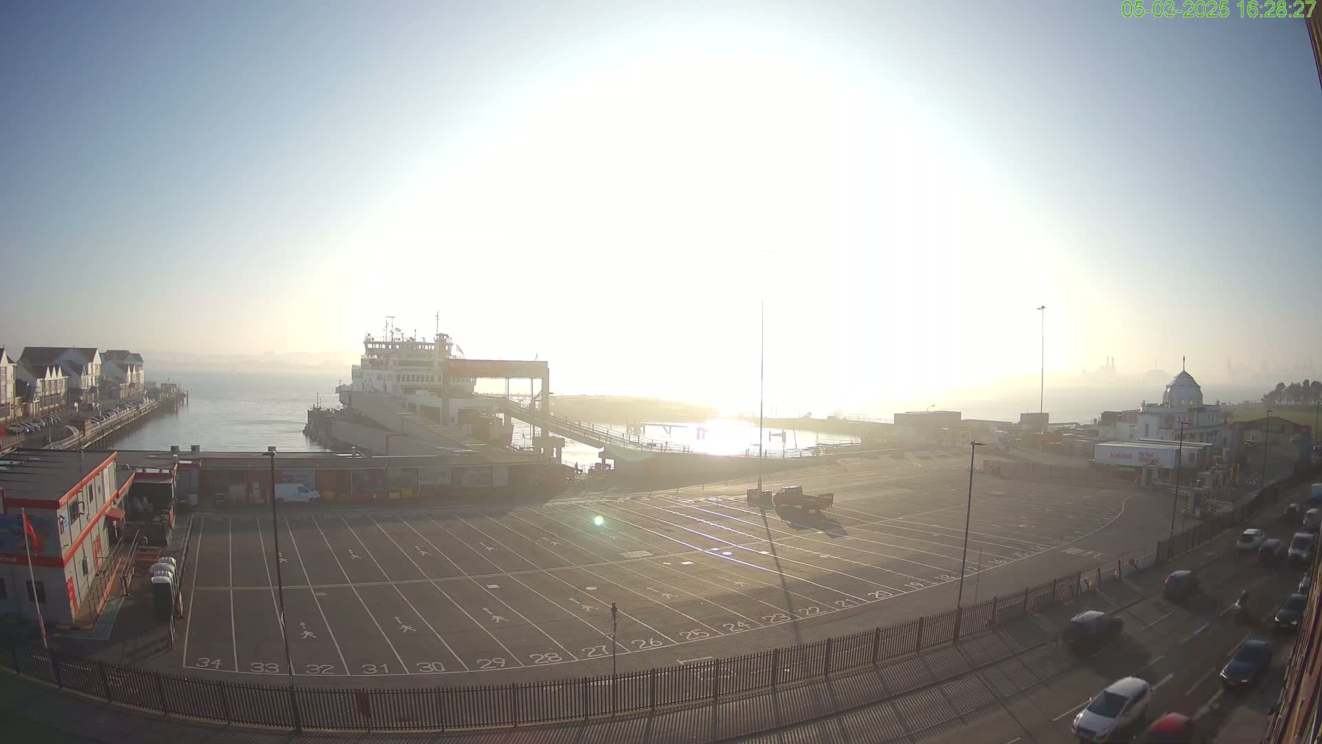 A ferry is docked at a terminal on a hazy, sunny morning, with cars driving on a road beside a large empty parking lot.