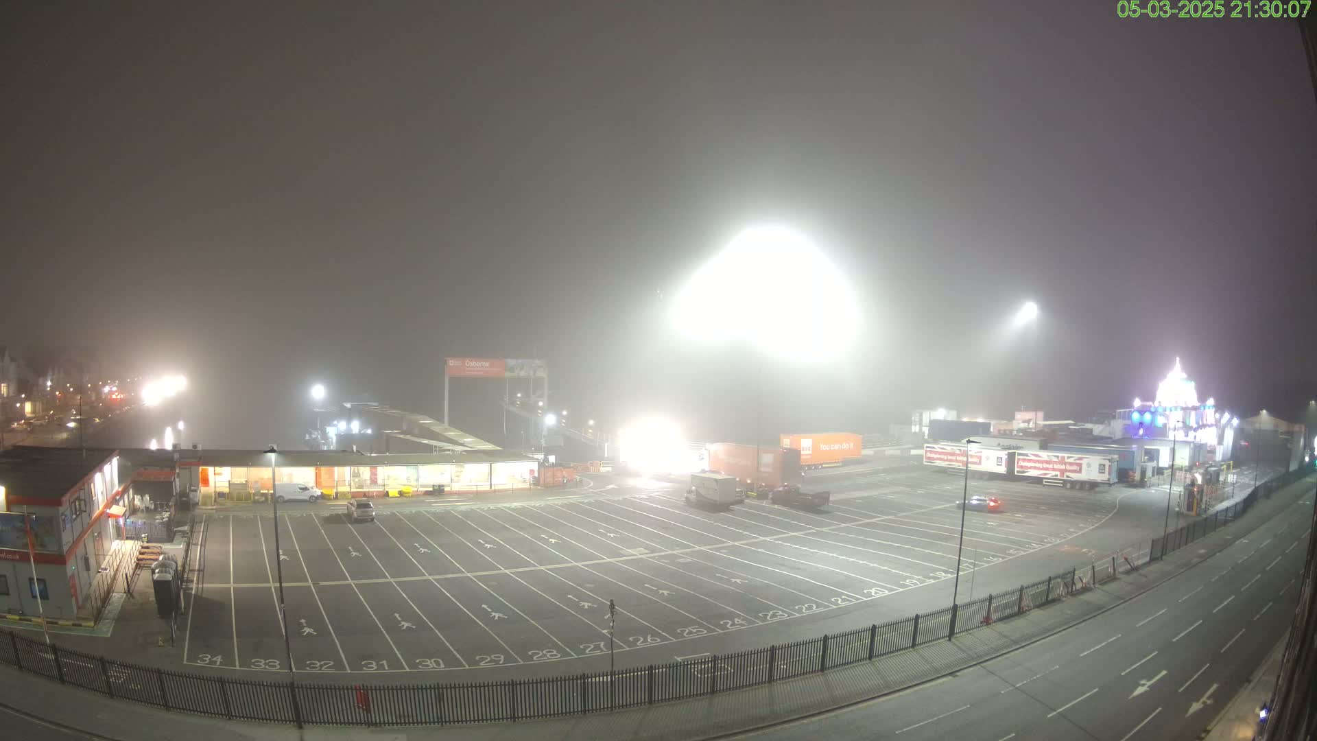 A foggy night scene shows an empty parking lot next to a ferry terminal with several semi-trucks parked nearby.