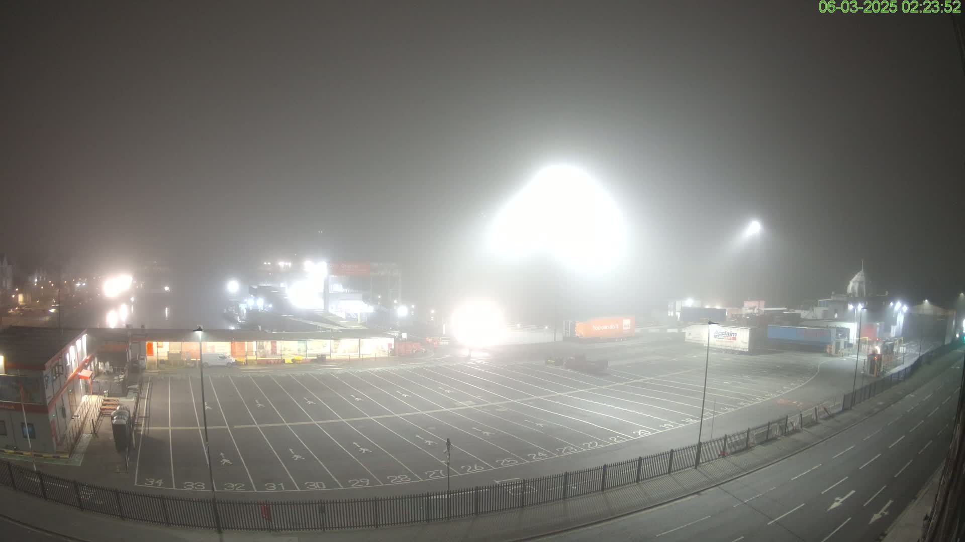 A foggy, brightly lit nighttime parking lot with buildings and trucks in the background.