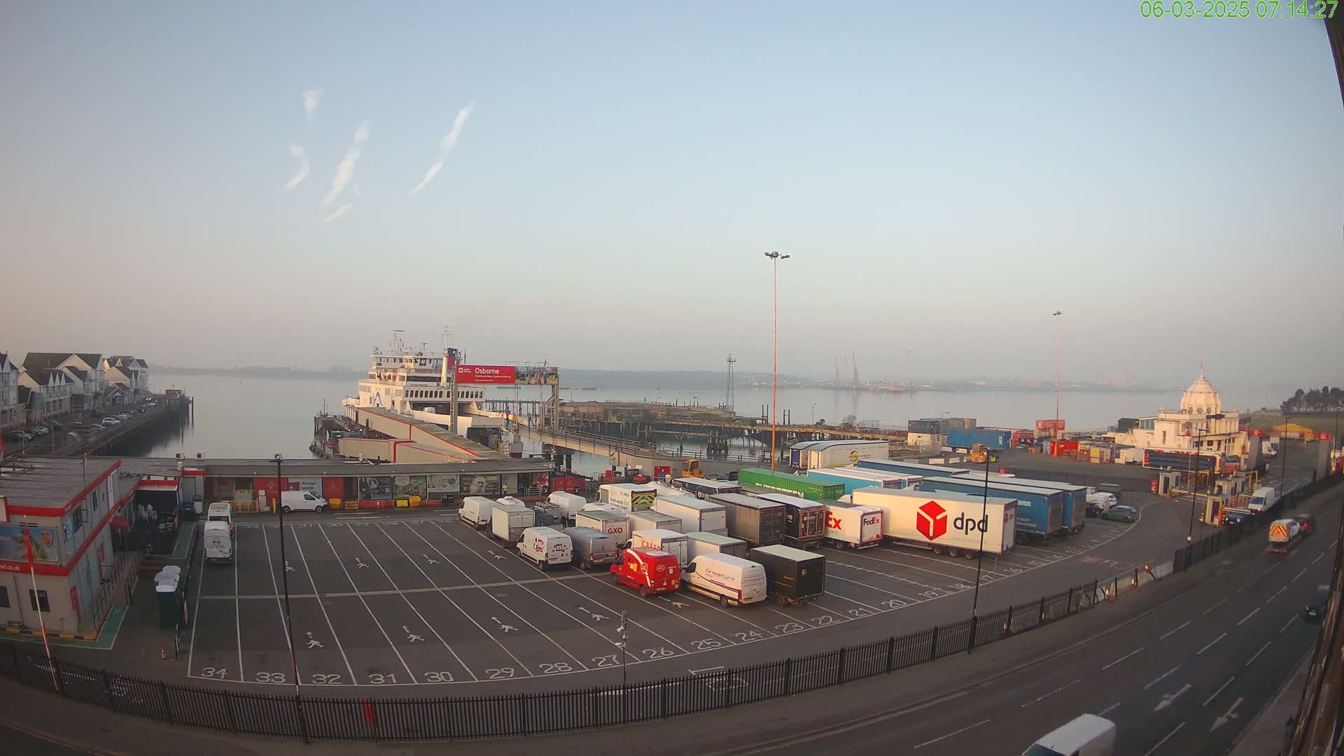 A ferry is docked at a port on a hazy morning, with numerous delivery trucks parked in a lot nearby.