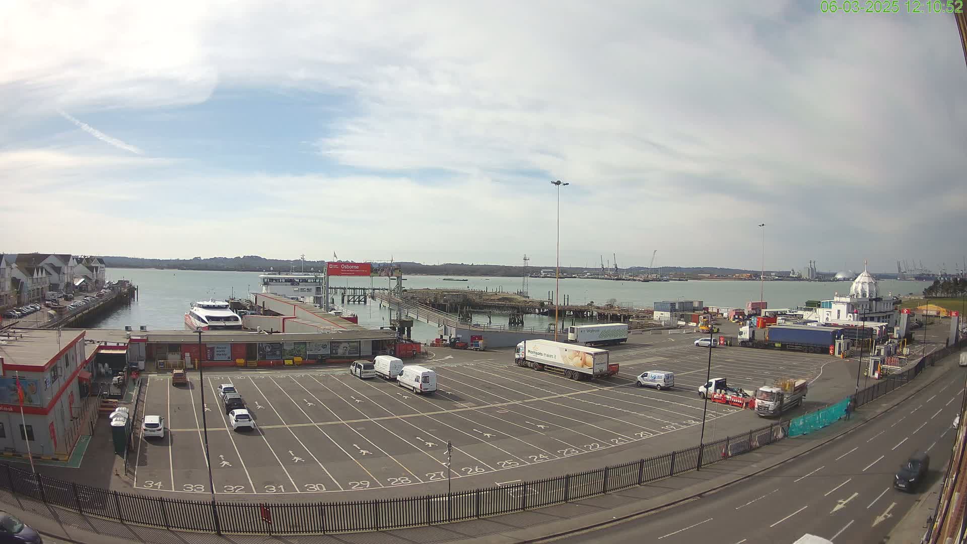 An empty parking lot at a ferry terminal overlooks a calm body of water under a partly cloudy sky.