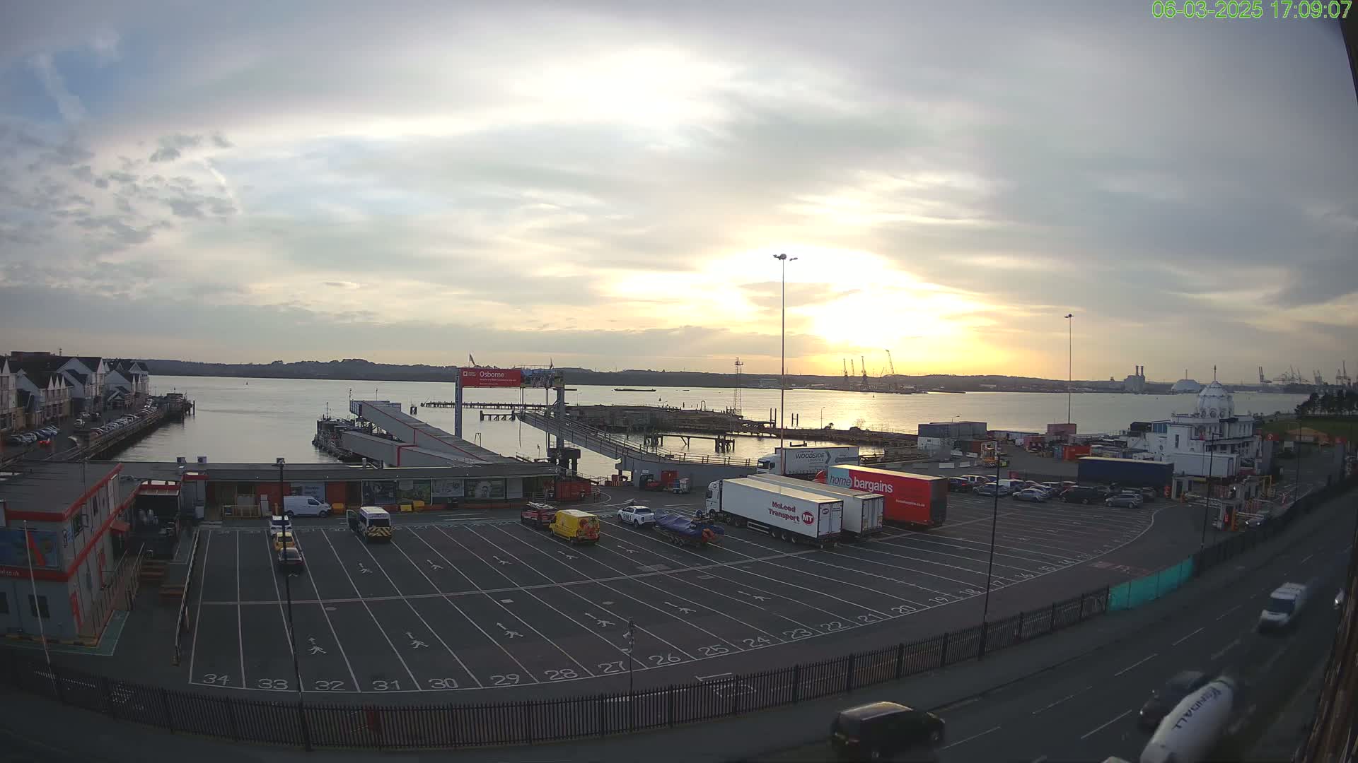 A sunset over a calm body of water, viewed from a high vantage point showing a ferry terminal with several vehicles parked in a large lot and additional vehicles driving along a road in front of the terminal.