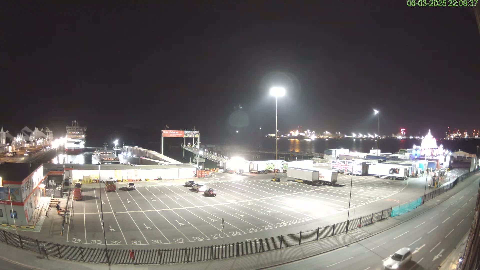 A nighttime view of a ferry terminal with several parked trucks and cars, a ferry docked at the pier, and a body of water in the background under a dark sky.