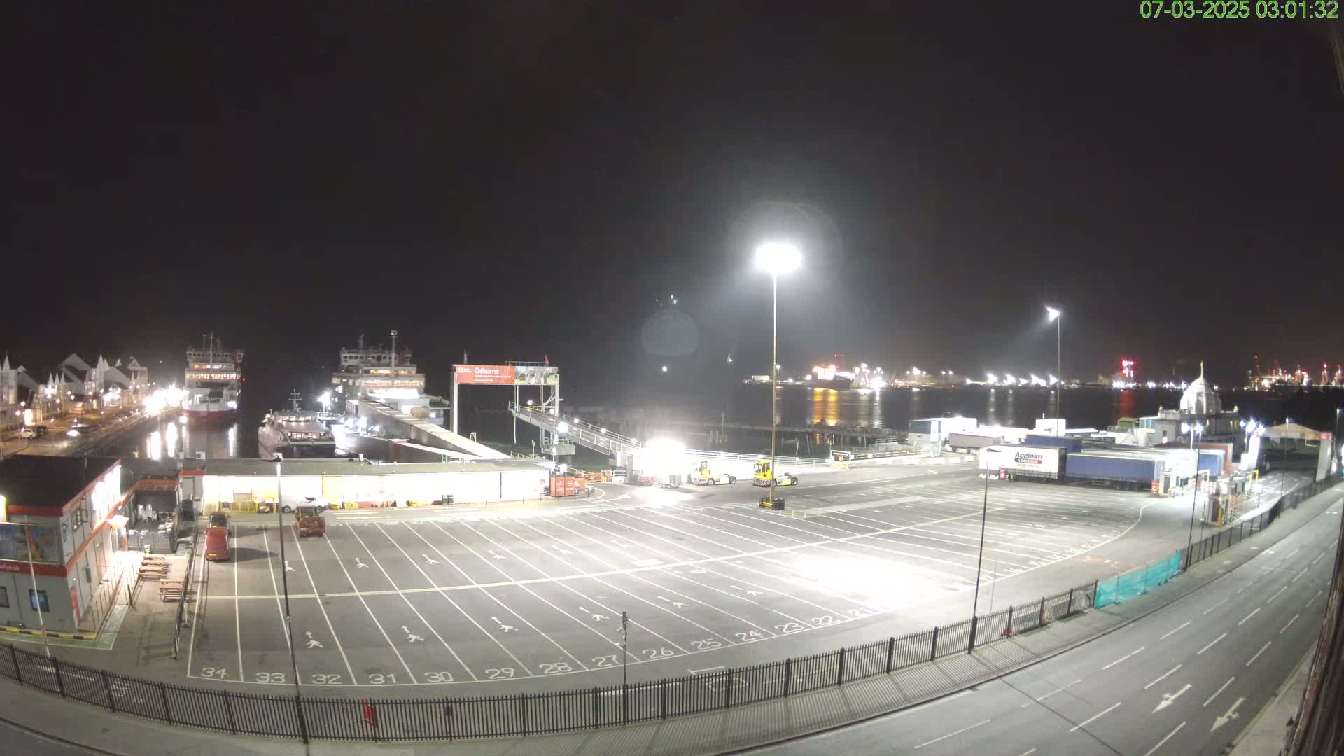 At night, a brightly lit ferry terminal shows two ferries docked, numerous parked trucks, and an empty parking lot next to a roadway.