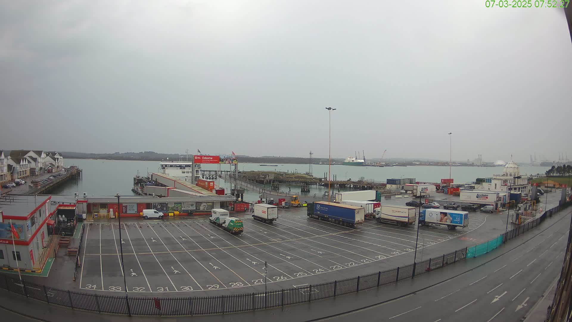 A ferry terminal parking lot with numerous trucks and a ferry in the background on a cloudy, overcast day.