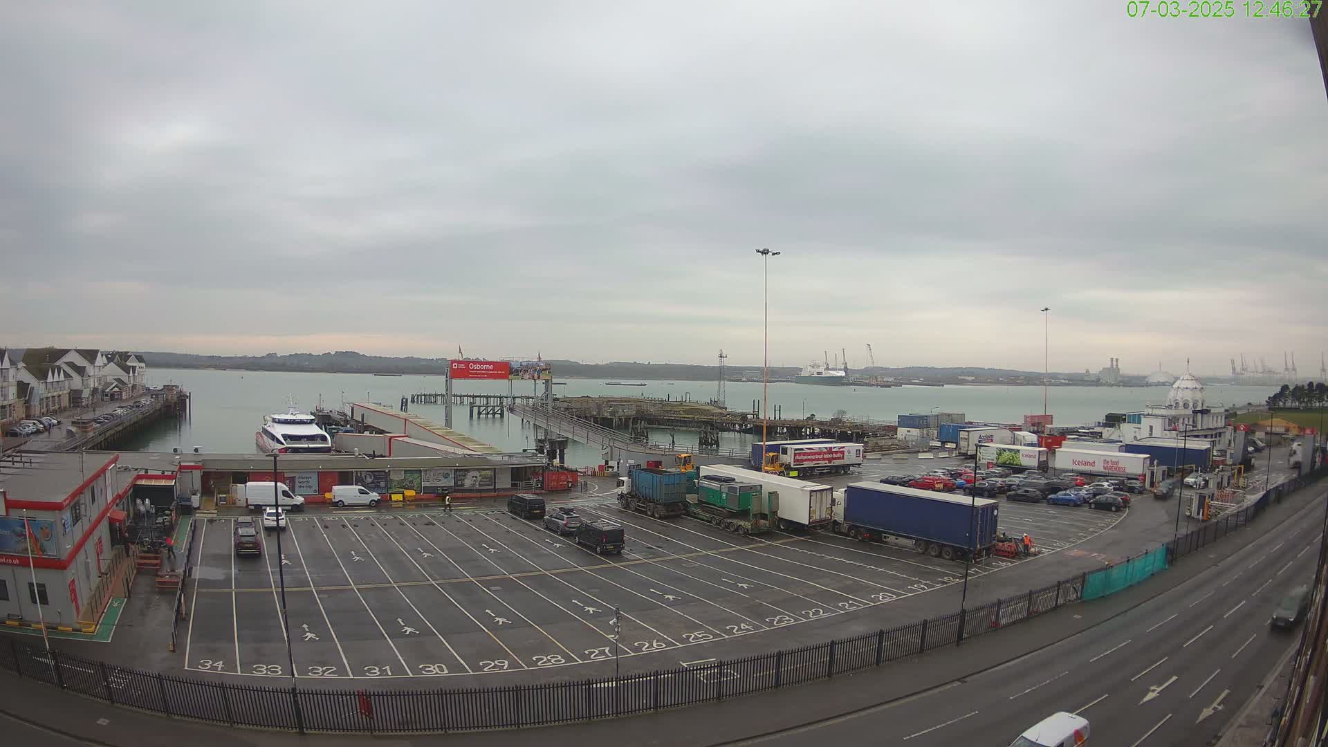 An overcast day at a ferry terminal shows a large parking lot filled with cars and trucks, a ferry docked at a pier, and calm water beyond.