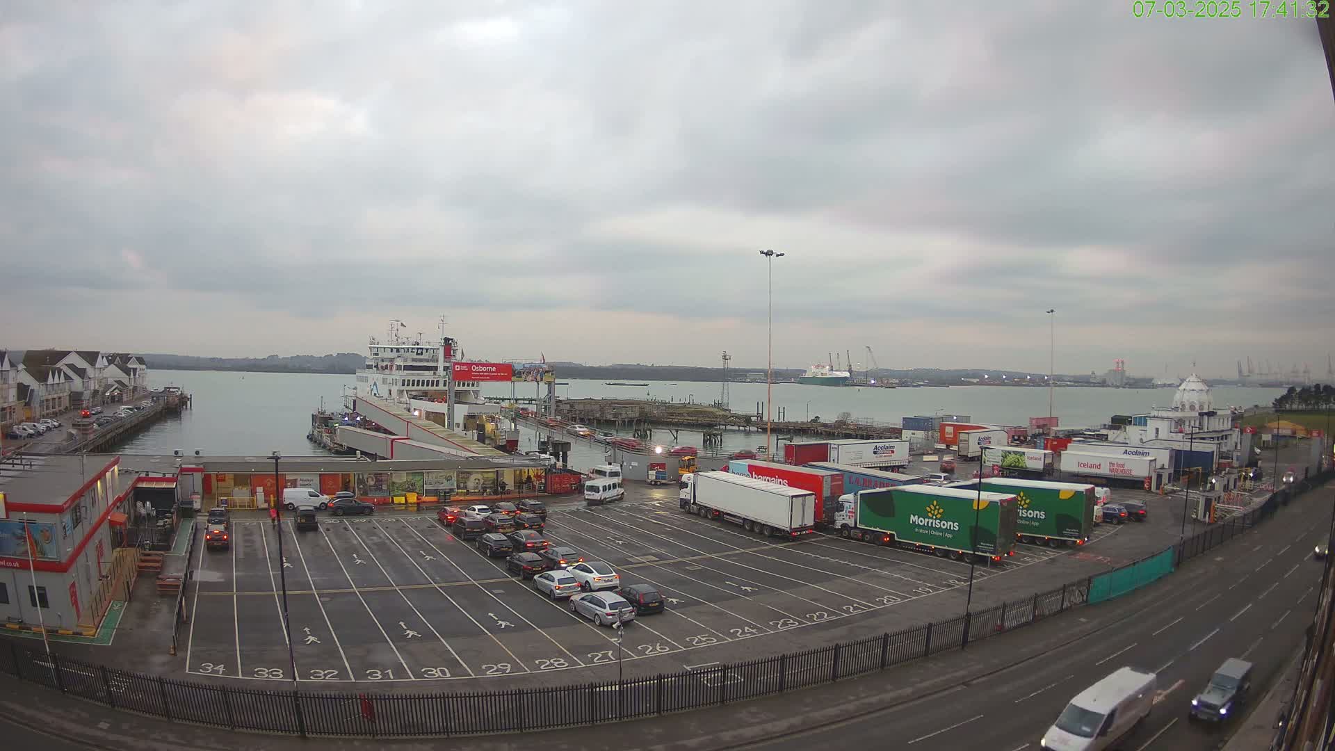 A ferry is docked at a port with numerous cars and trucks parked in a large lot under an overcast sky.