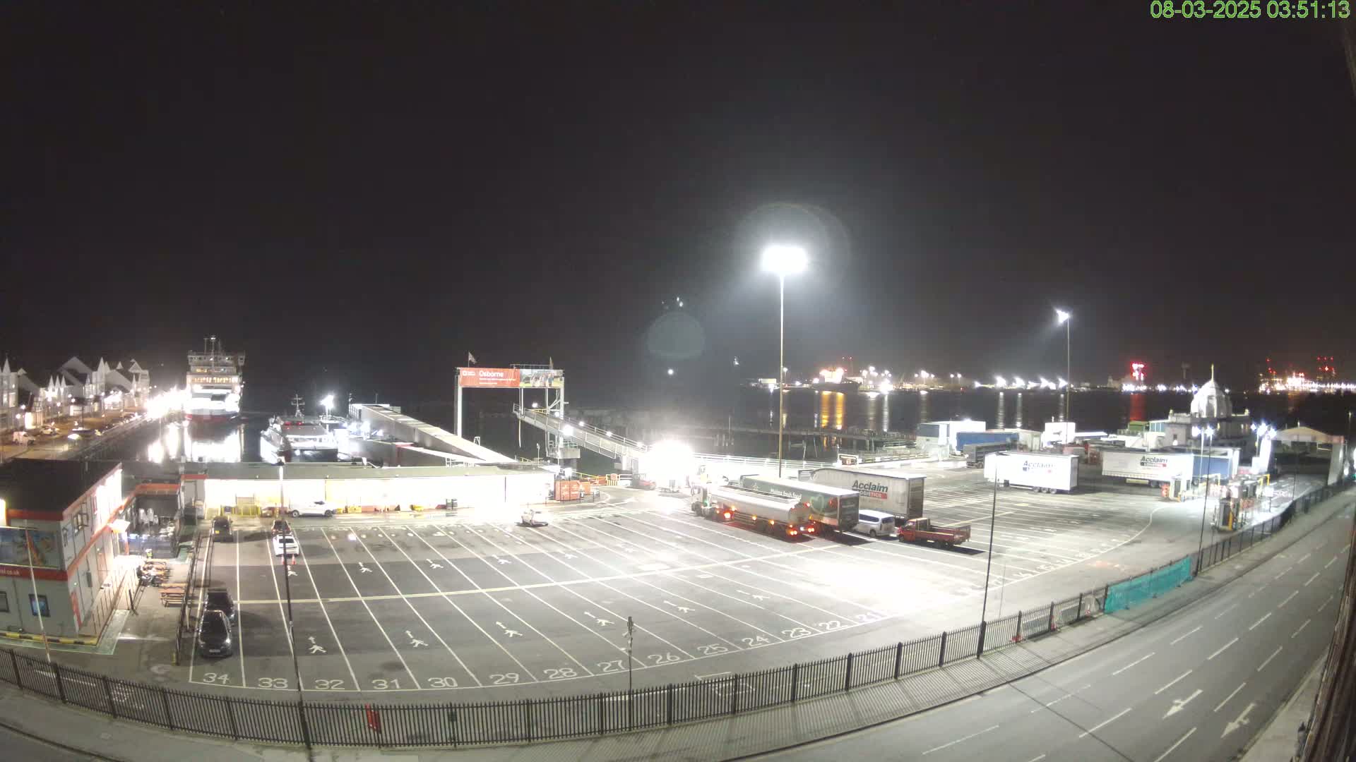A nighttime view of a ferry terminal with docked ferries, several parked trucks, and an empty parking lot illuminated by bright lights.
