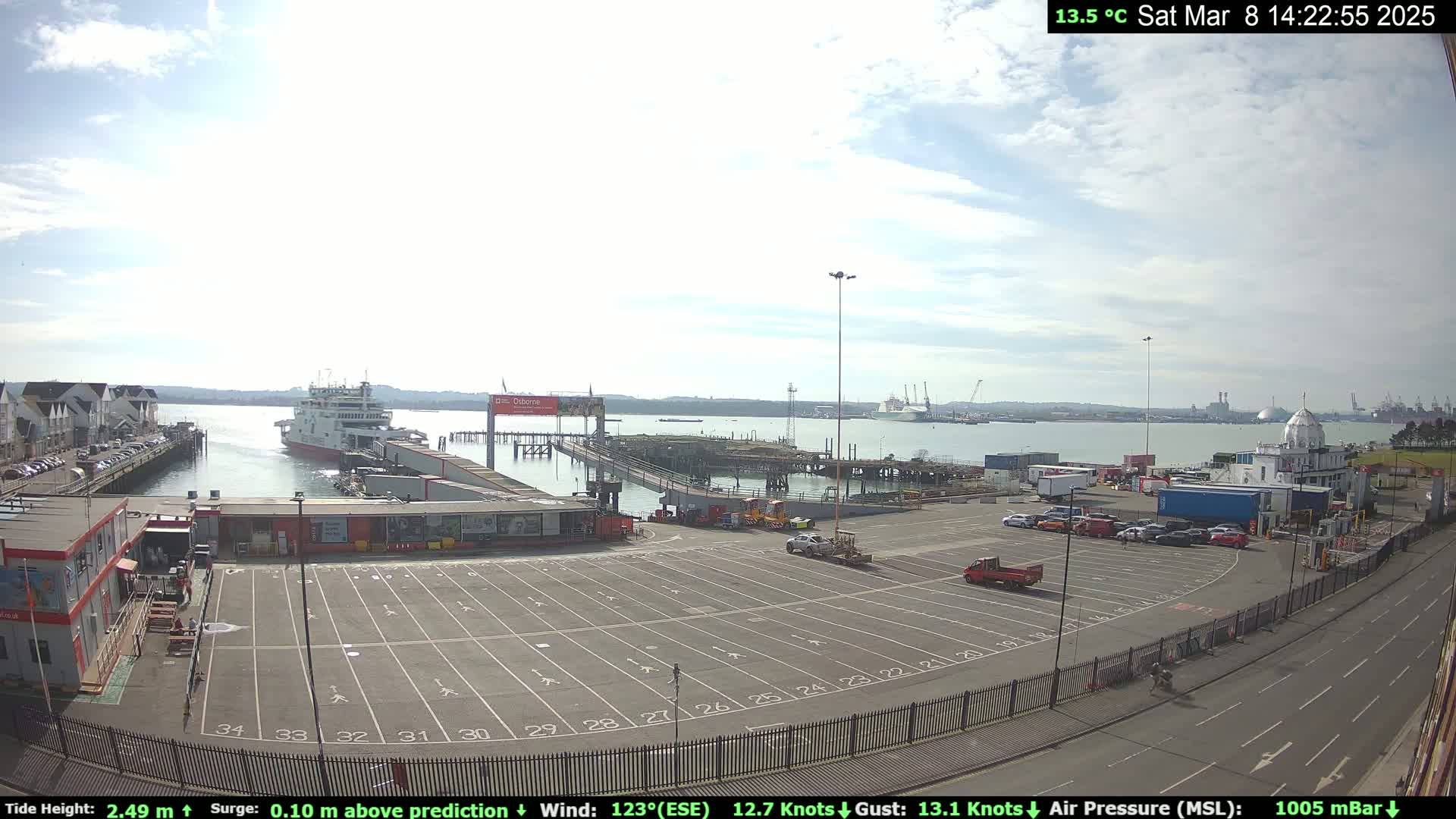 A ferry is docked at a port on a partly cloudy day, with a large empty parking lot and other vehicles visible nearby.