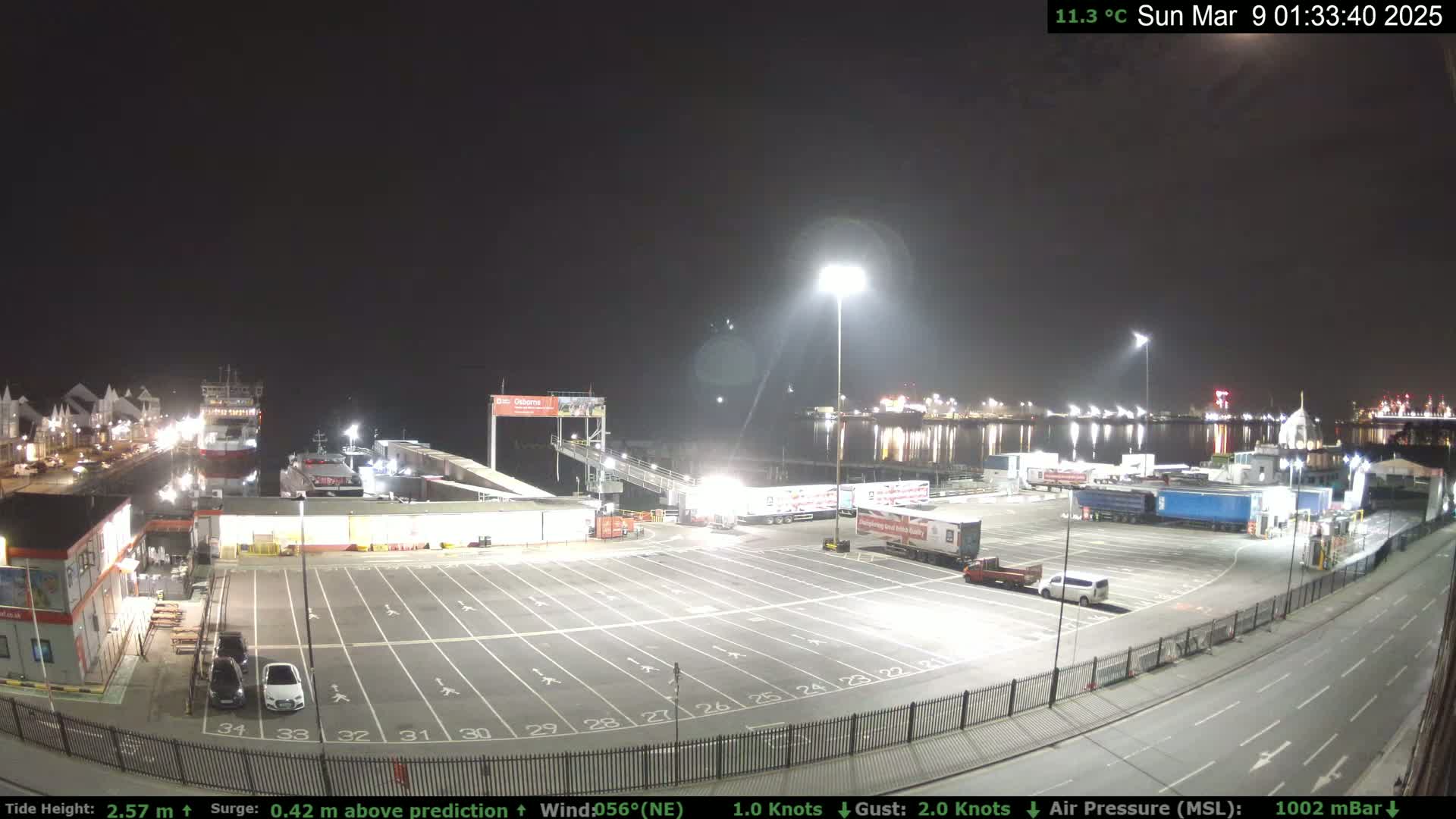 A nighttime view of a ferry terminal with several docked ferries, numerous trucks parked in a large lot, and cars in a smaller adjacent lot; the weather is clear at 11.3 degrees Celsius.