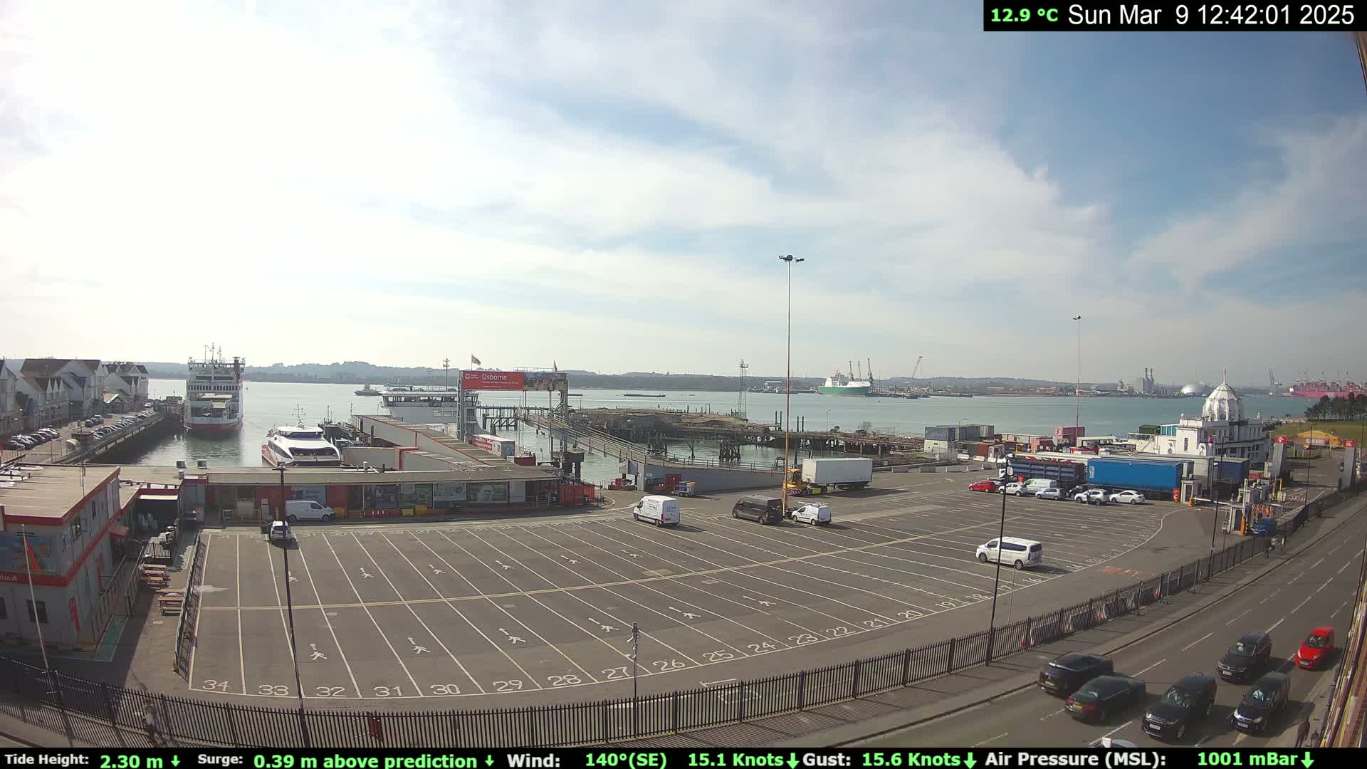 A mostly sunny day at a ferry terminal shows a large empty parking lot, several cars and vans, and a ferry docked at a pier, with additional ships visible in the distance.