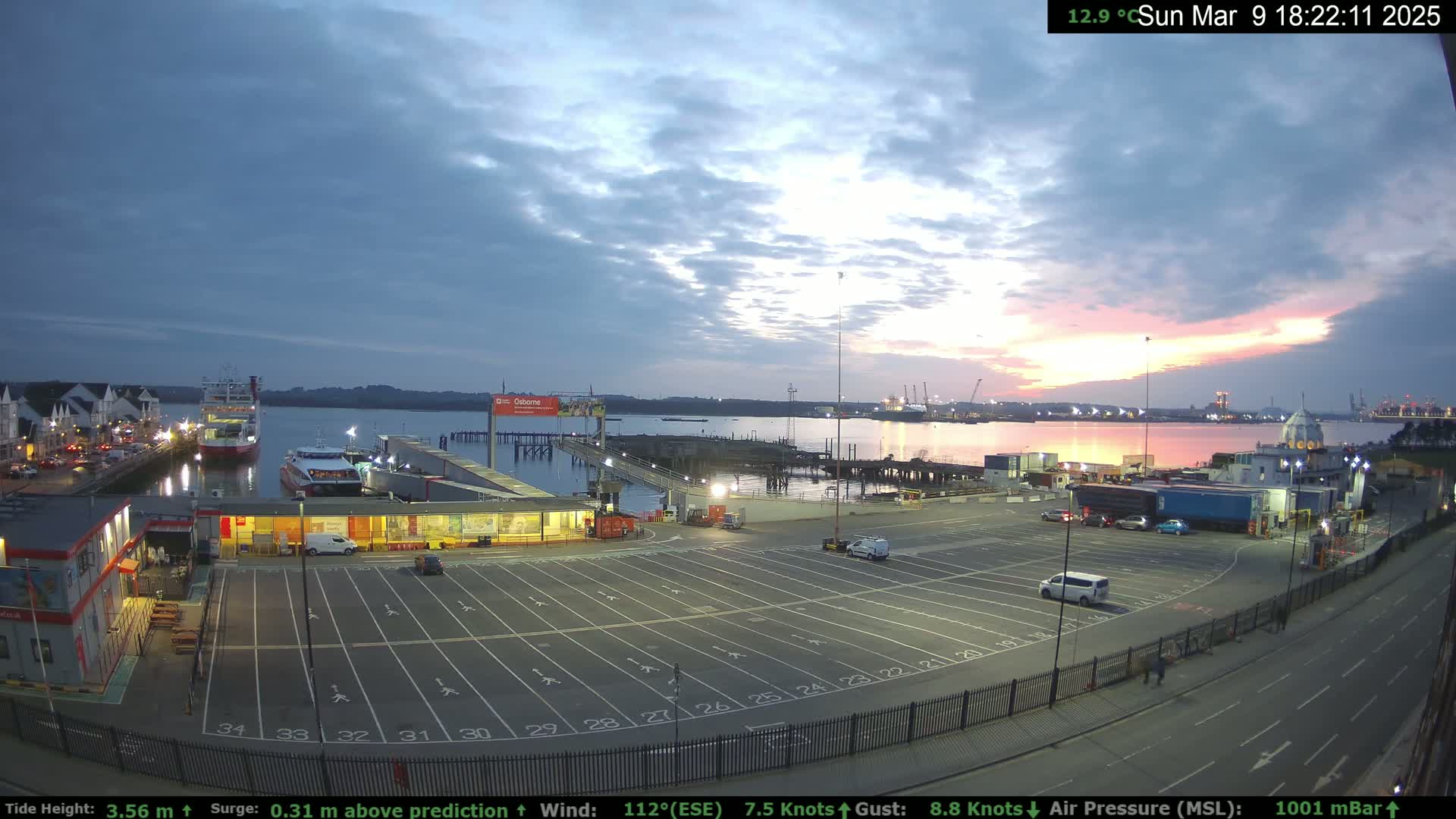 An empty ferry terminal parking lot at sunset under a partly cloudy sky, with a ferry and other boats visible in the water.