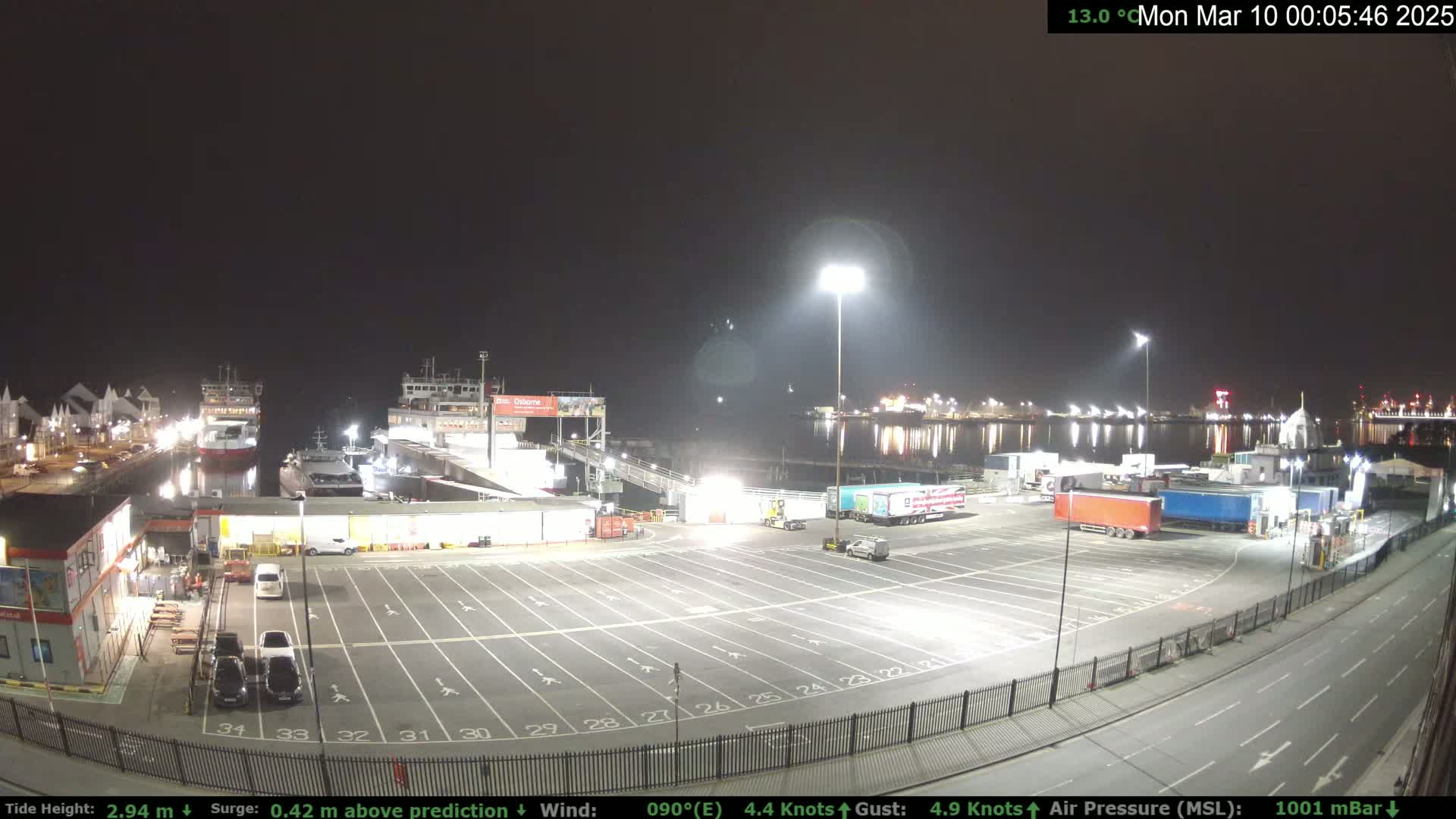 A nighttime view of a ferry terminal with several docked ferries, a large empty parking lot, and several semi-trailer trucks.