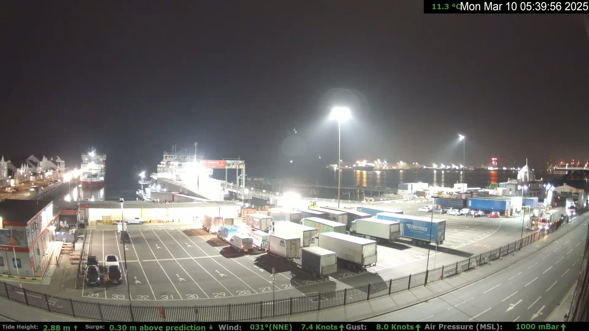 A nighttime view of a ferry terminal with several ships docked, numerous trucks parked in a lot, and cars on the road, under clear skies.