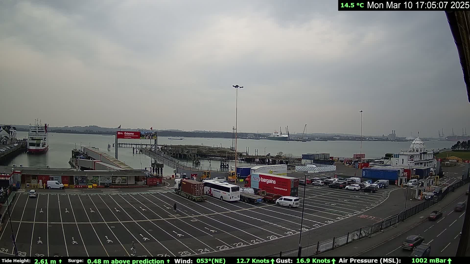 A ferry terminal with numerous vehicles parked in the lot, a ferry in the water, and overcast skies.