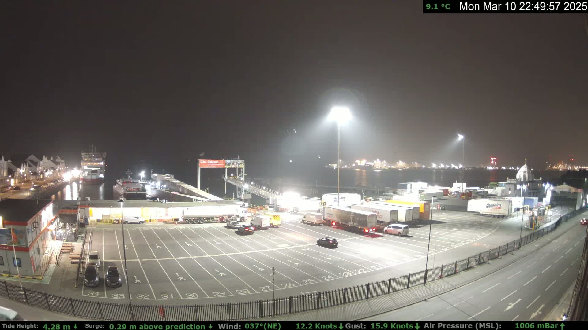 A nighttime view of a ferry terminal with several trucks and cars parked in a lot, under a dark, hazy sky.