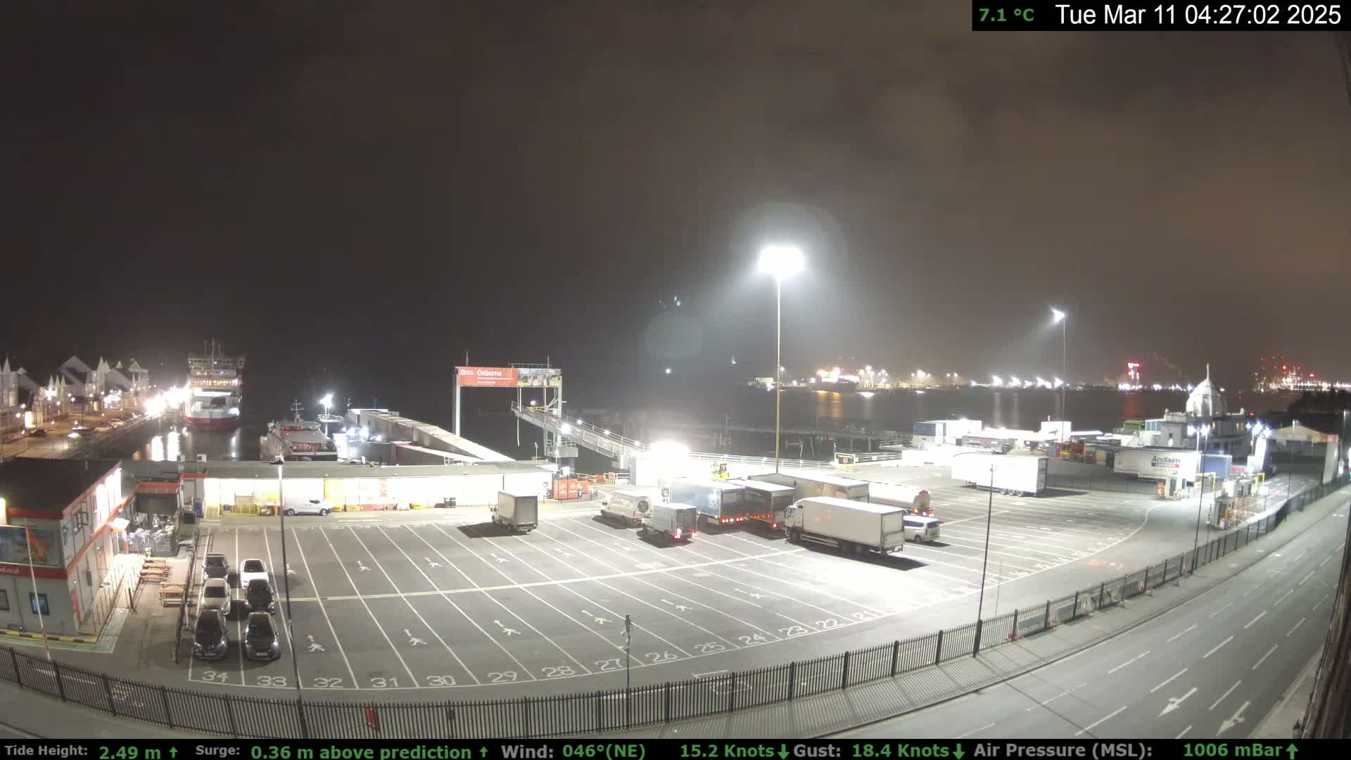 A nighttime view of a ferry terminal with several trucks and cars parked in a lot, a ferry docked at the pier, and calm water in the background.