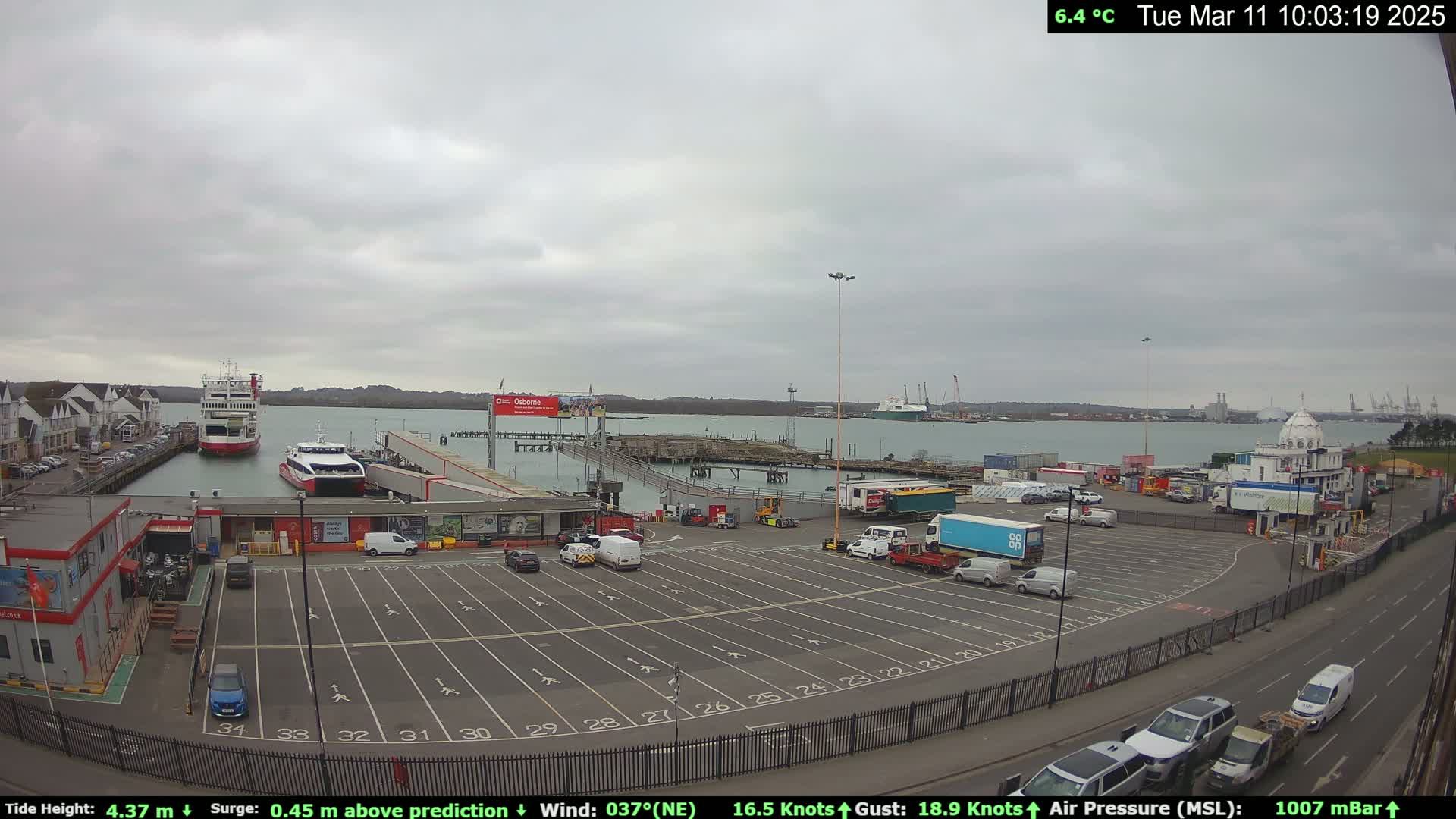An overcast day at a ferry terminal shows several vehicles parked in a lot, with two ferries docked and additional watercraft in the harbor.