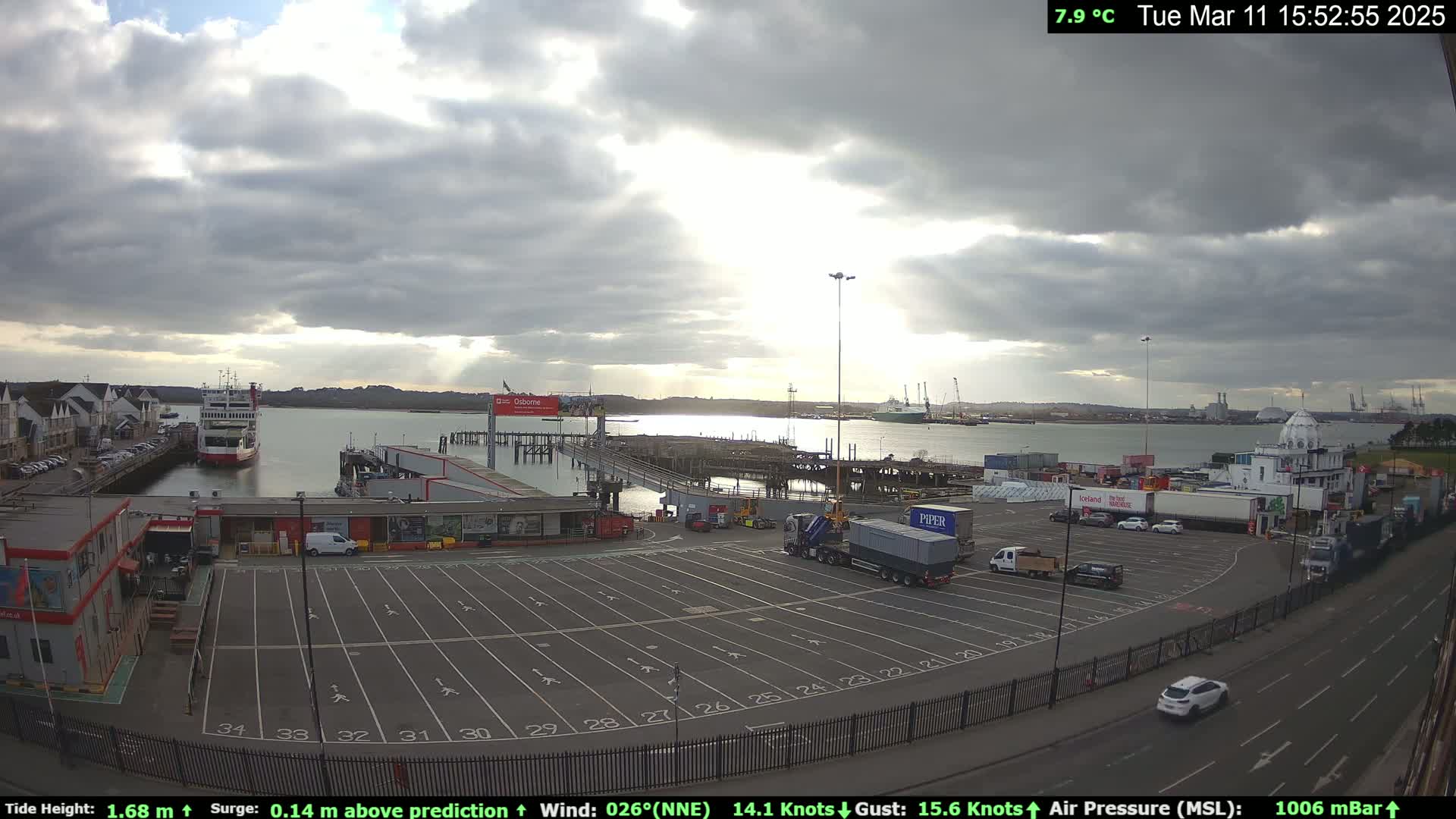 A ferry is docked at a port under a partly cloudy sky, with a large empty parking lot and several vehicles visible.