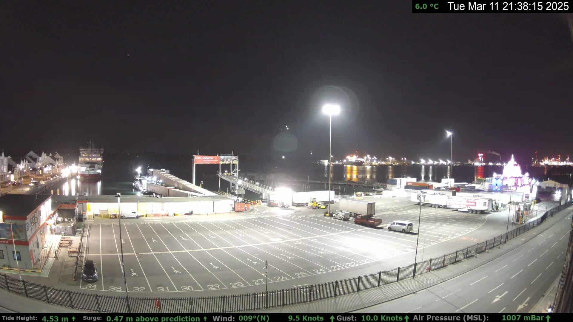 A nighttime view of a ferry terminal with a large empty parking lot, several trucks, and a ferry docked; the weather is clear and cool at 6.0°C.