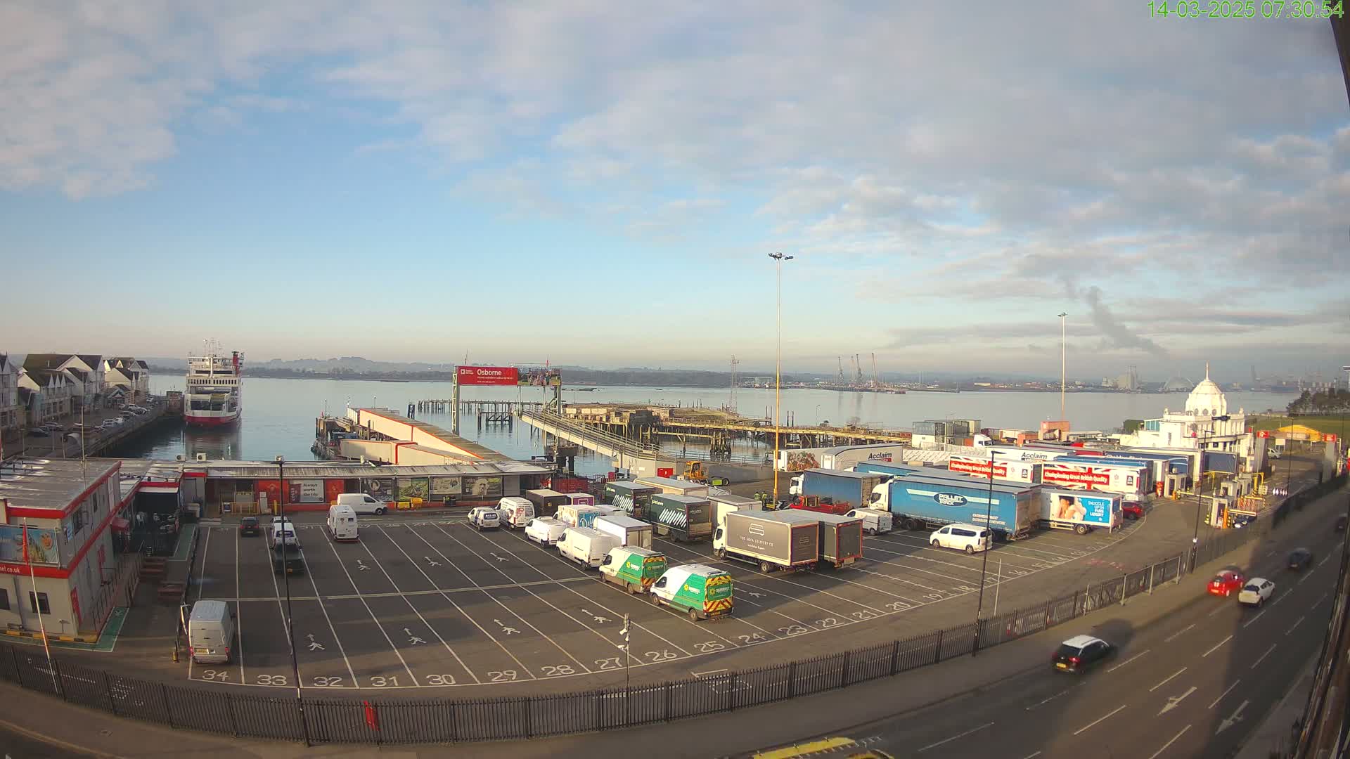 A ferry is docked at a port with numerous trucks and vans parked in a lot, under a partly cloudy sky.