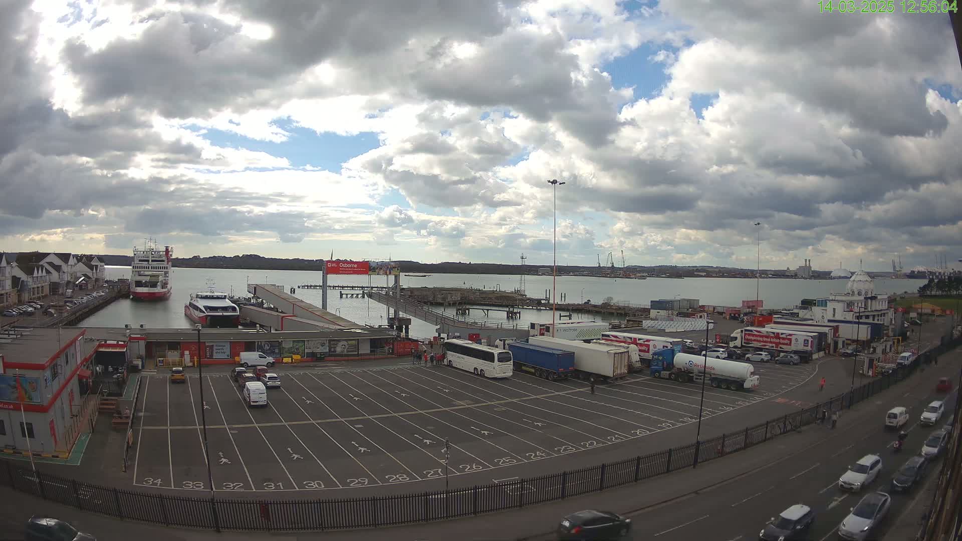 A ferry terminal with two ferries docked, numerous vehicles parked, and a partly cloudy sky.