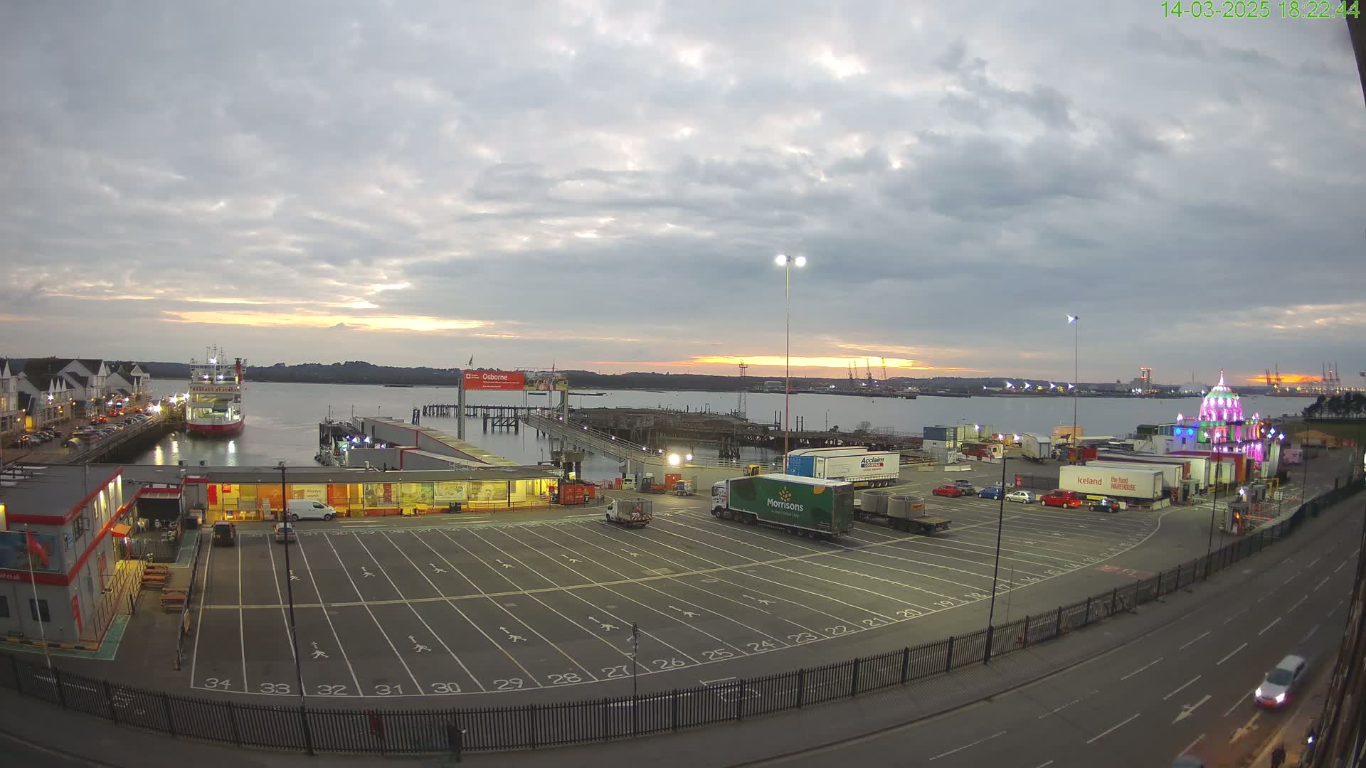A mostly cloudy sunset over a harbor, with a ferry docked and numerous trucks parked in a lot adjacent to a roadway.
