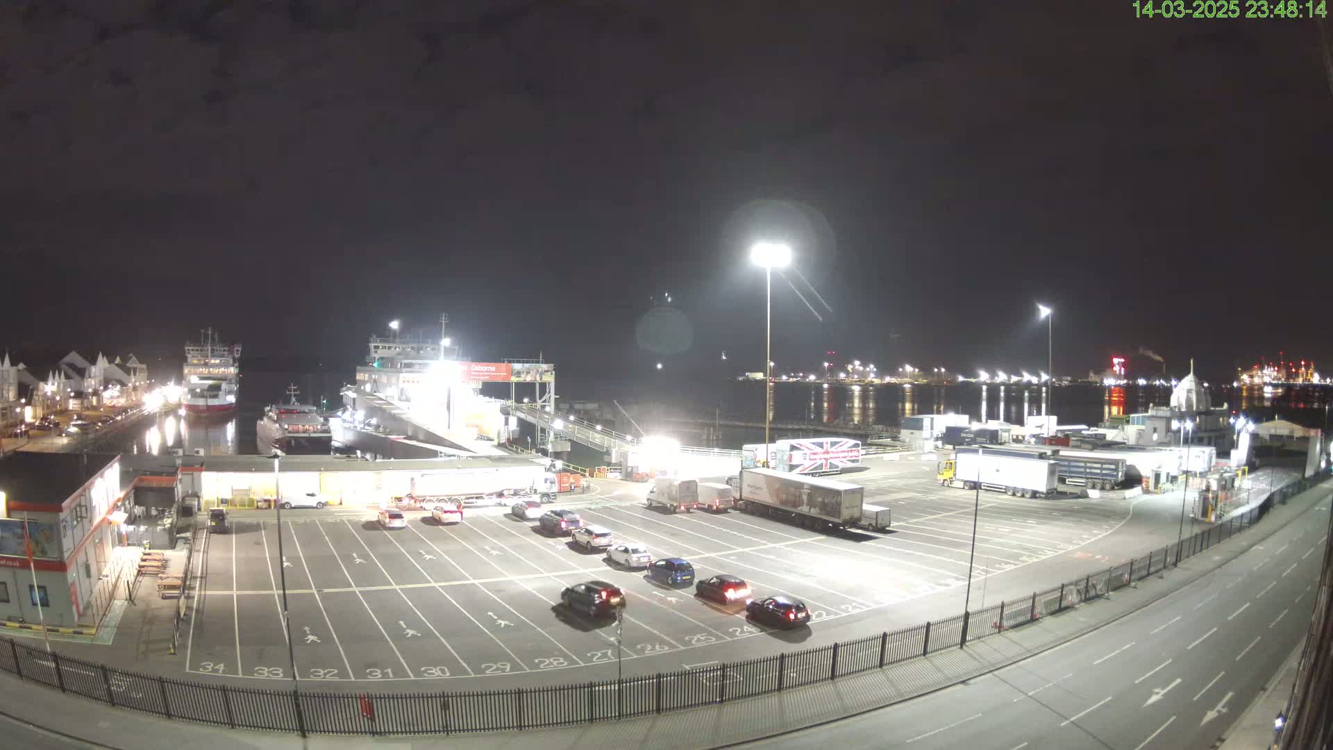 A nighttime view of a ferry terminal with several cars parked in a lot, two ferries docked, and numerous large trucks in the background, under a clear dark sky.