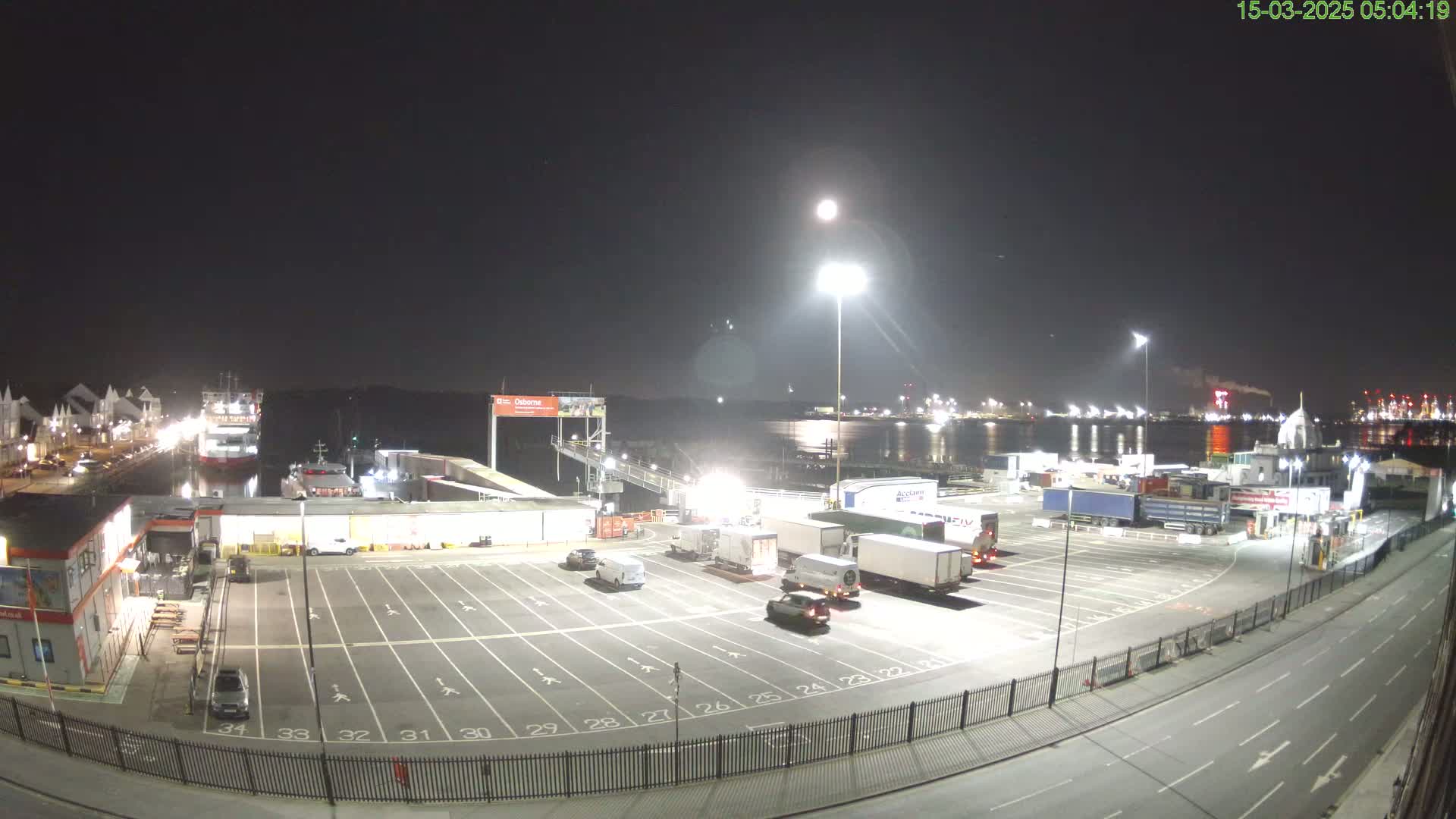 A nighttime view of a ferry terminal with several trucks parked in a lot, a ferry docked, and a body of water in the background under a bright moon.