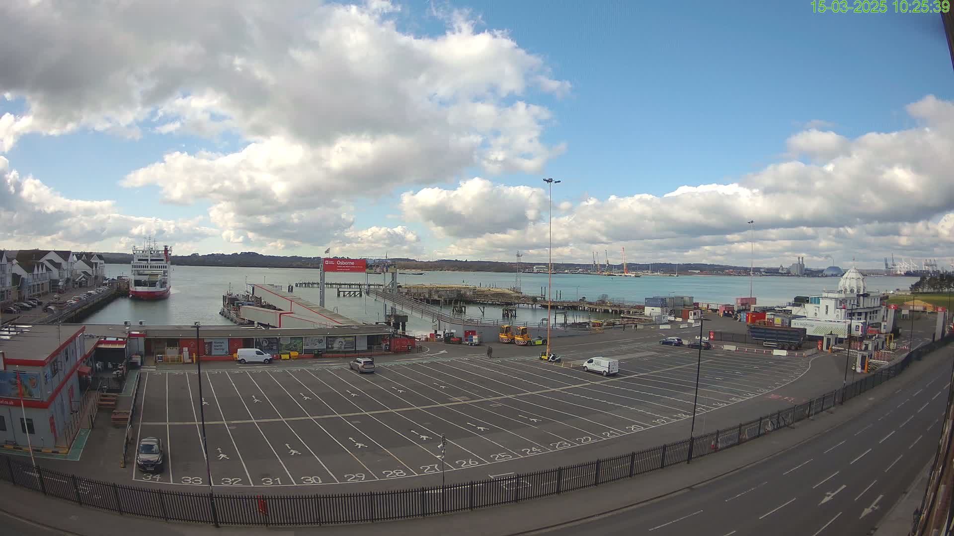 A ferry is docked at a port under a partly cloudy sky, with a large, mostly empty parking lot and a roadway visible in the foreground.