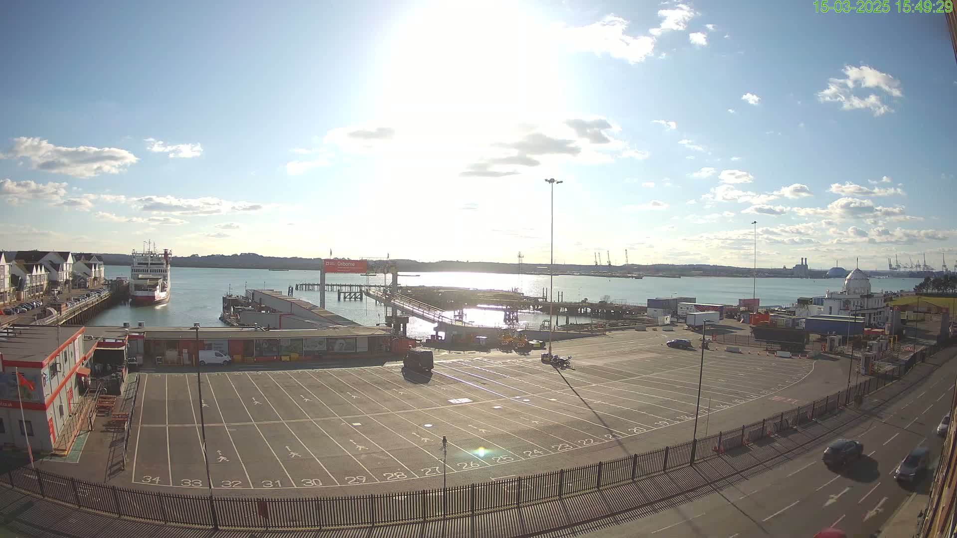 A ferry is docked at a port on a sunny day with some clouds, and a large empty parking lot is visible, along with roads and other buildings.