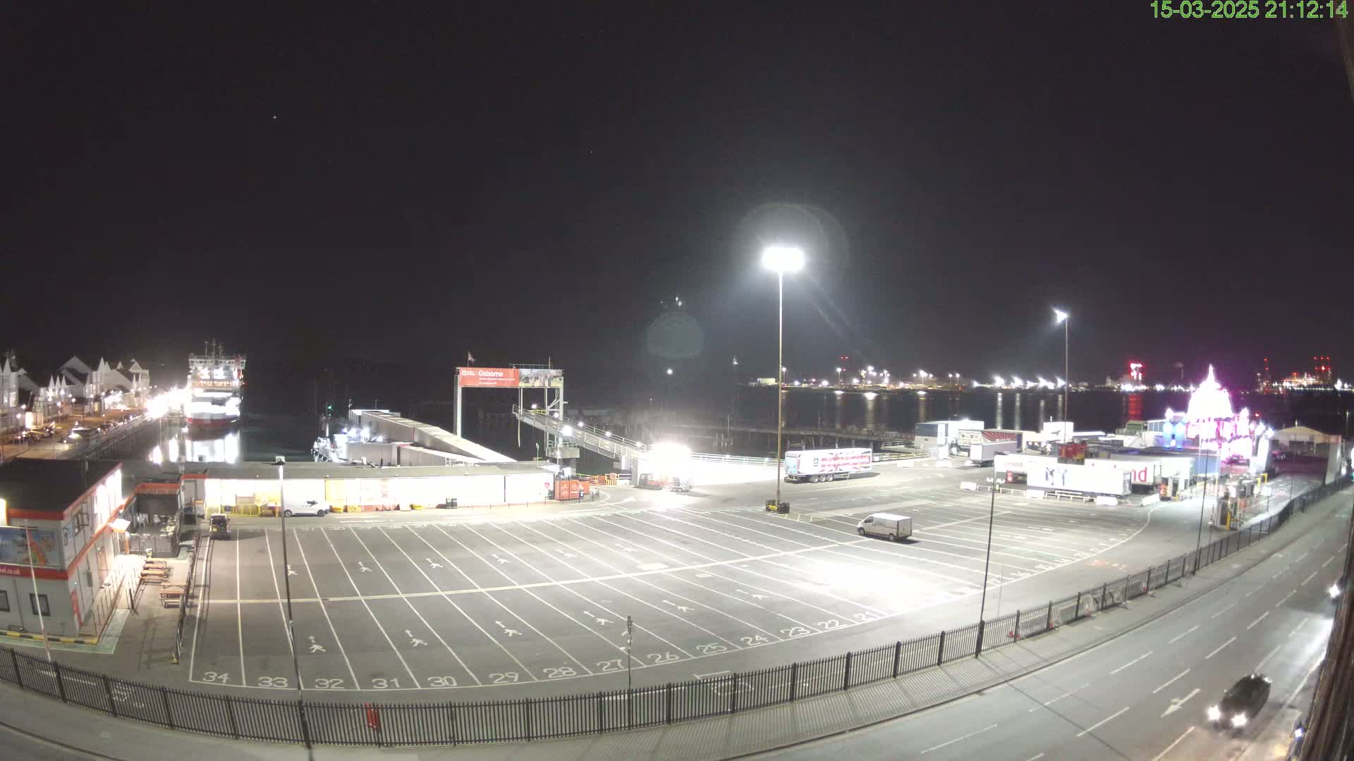 A nighttime view of a ferry terminal with a large empty parking lot, a docked ferry, and several trucks and cars.