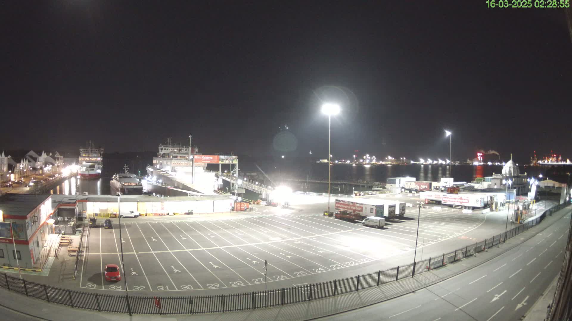 A nighttime view of a ferry terminal with several ships docked, a large empty parking lot, and trucks parked nearby, under clear skies.
