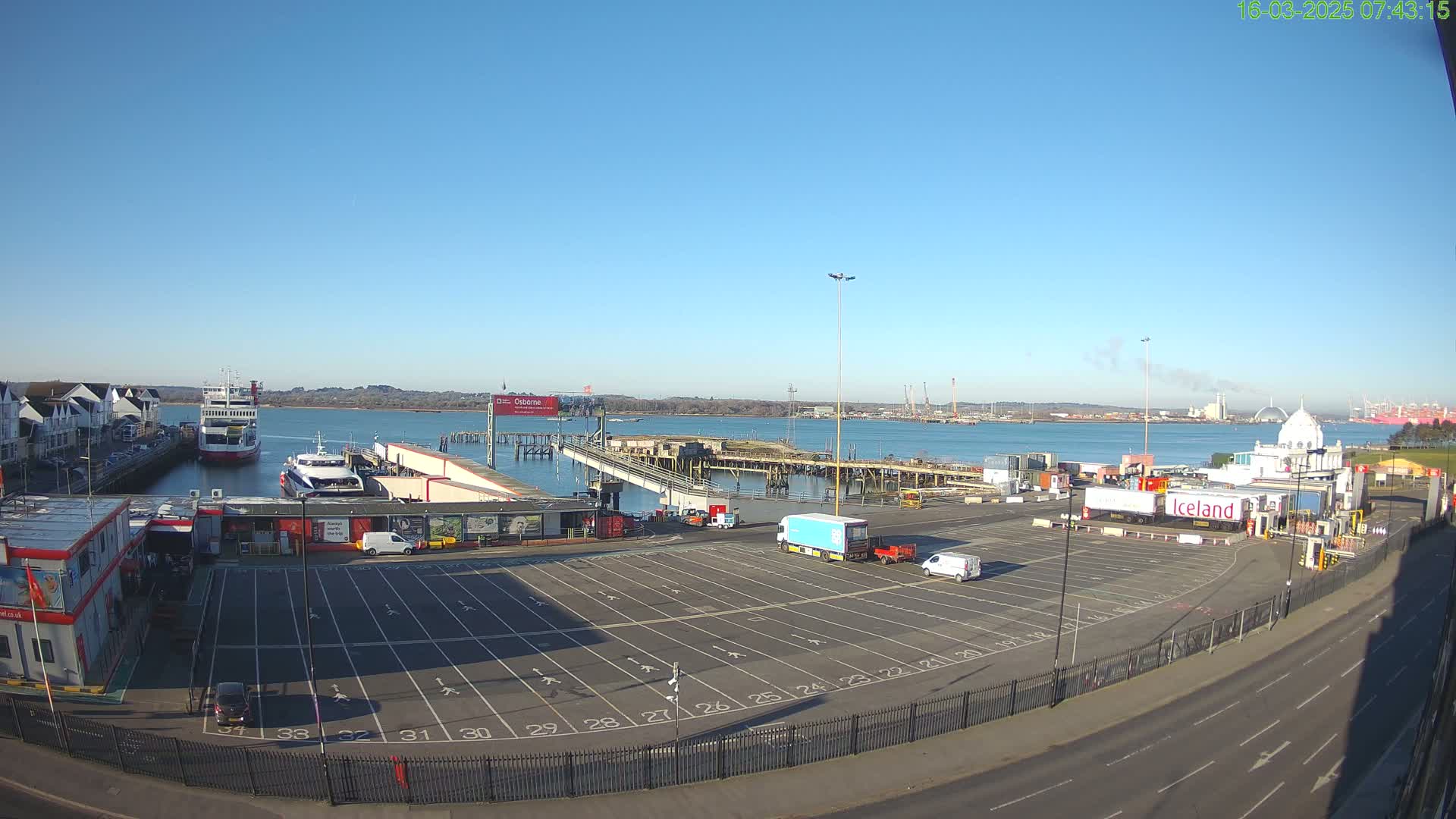 A ferry terminal with a large empty parking lot, a ferry boat docked, and calm waters under a clear blue sky.