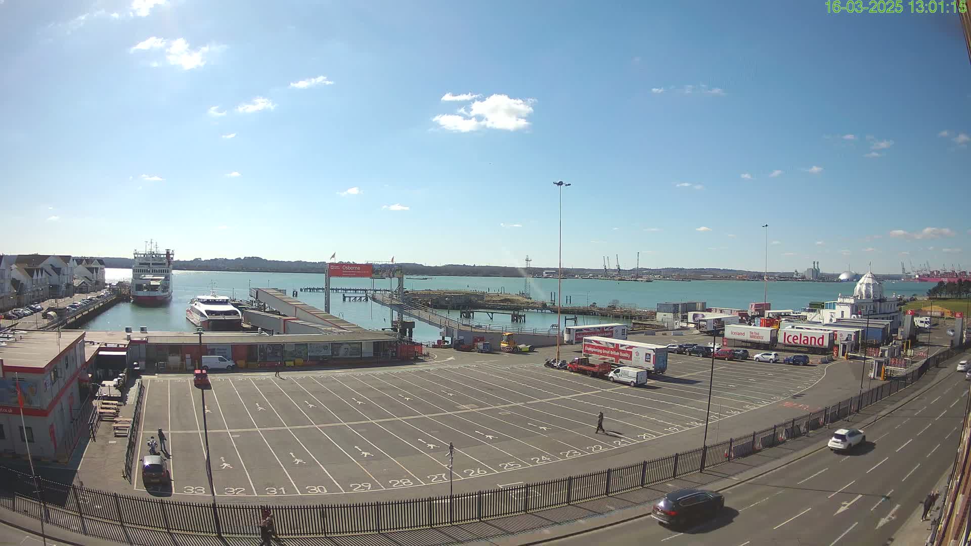 A ferry terminal with a large empty parking lot, several vehicles, and two ferries in the water under a mostly sunny sky.