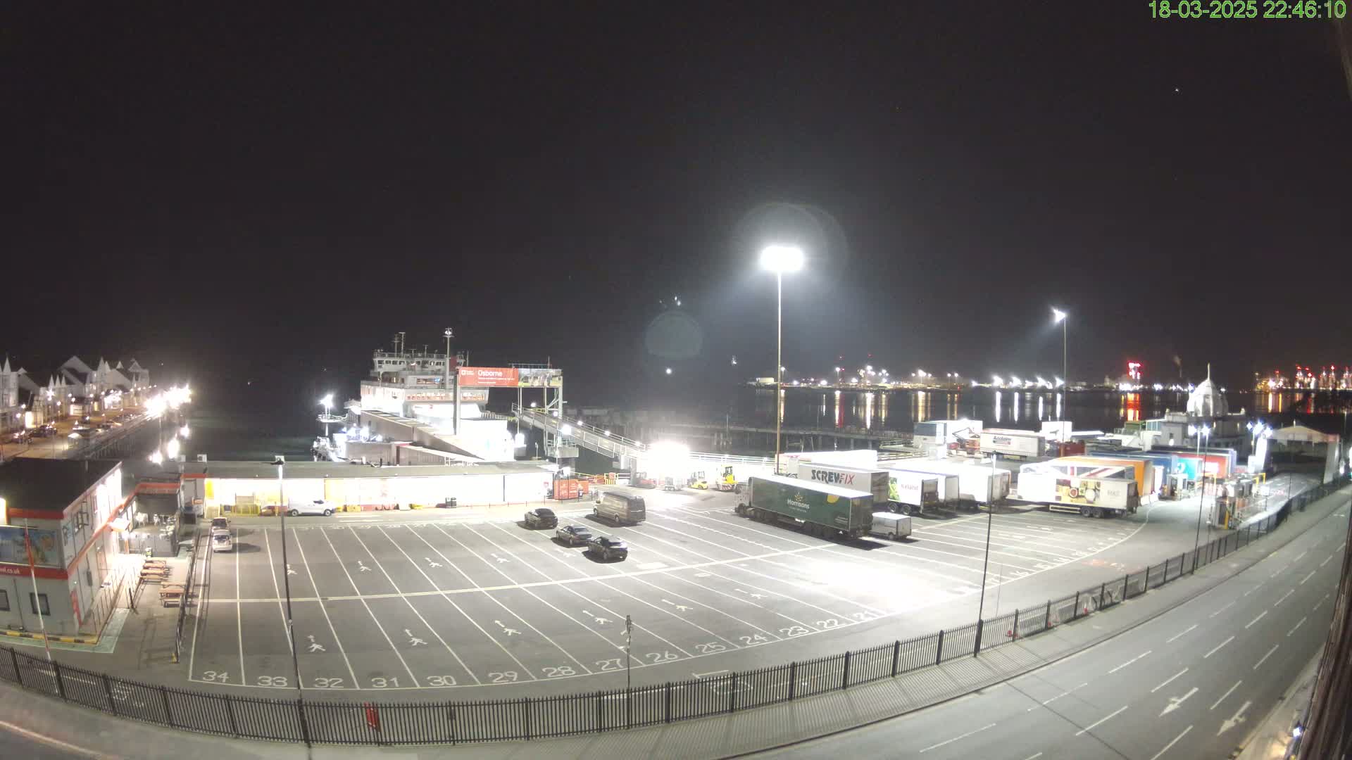 A nighttime, brightly lit ferry terminal with several cars and large trucks parked in a lot, a ferry docked, and a calm body of water in the background.