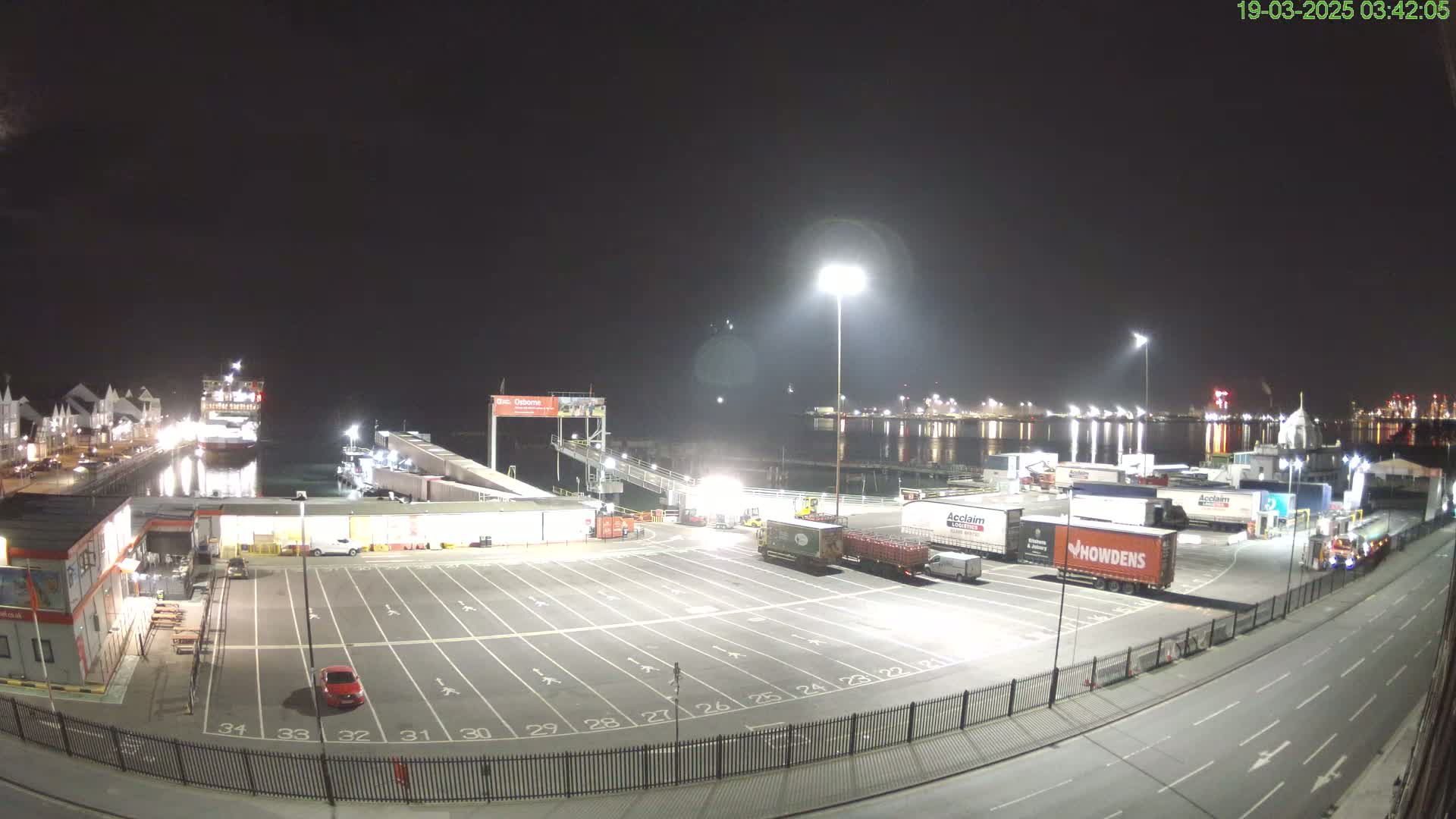 A nighttime view of a ferry terminal with a ferry docked, numerous trucks parked in a lot, and a dark, calm body of water reflecting lights in the background.
