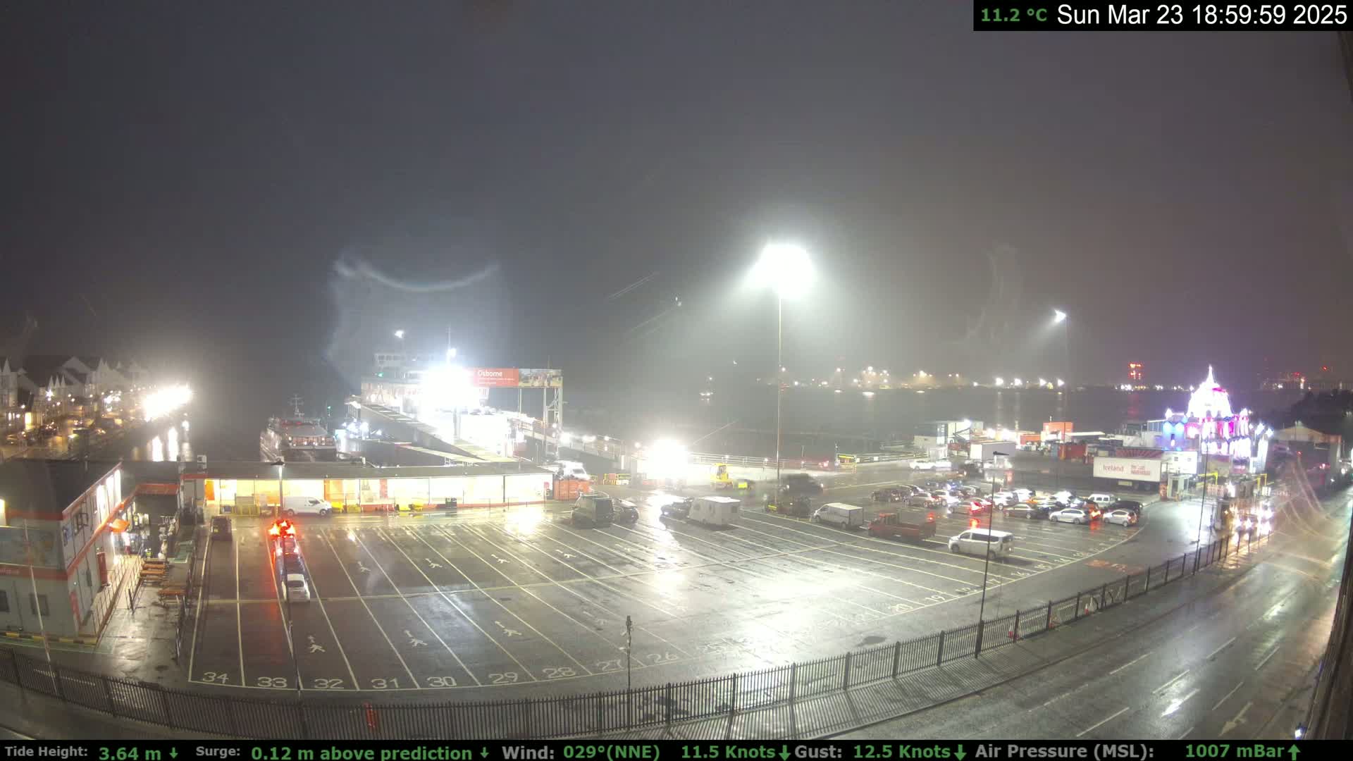A nighttime view of a ferry terminal with rain, showing a mostly empty parking lot, several vehicles, and a ferry in the background.