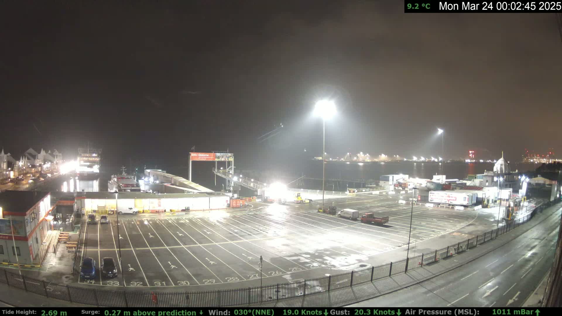 A nighttime view of a ferry terminal with a mostly empty parking lot, several vehicles, and a ferry docked, under overcast skies with strong winds.