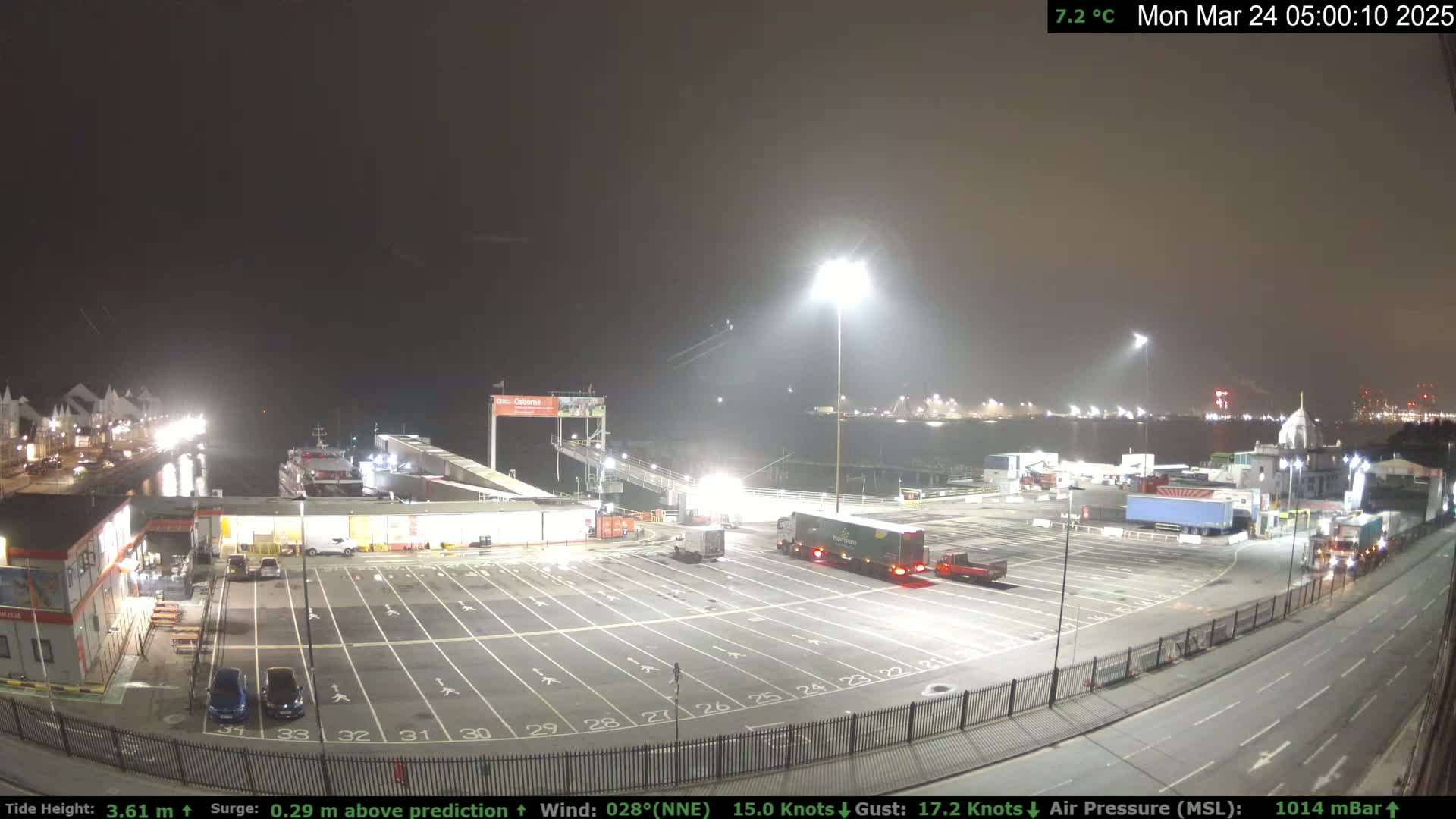 A nighttime view of a ferry terminal with several trucks and cars in a parking lot, under hazy conditions and a 15-knot wind.