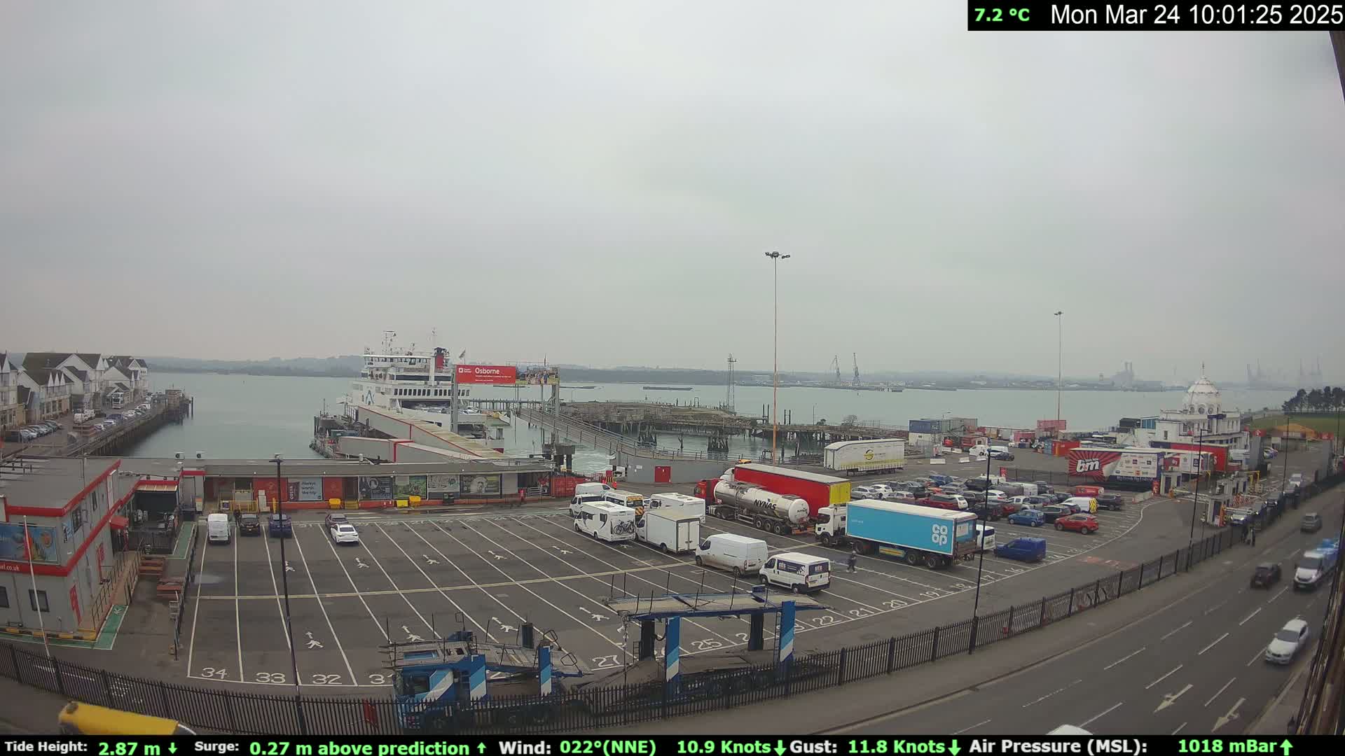 A ferry is docked at a port on an overcast day, with numerous vehicles parked in the surrounding lot and additional vehicles driving on the road nearby.