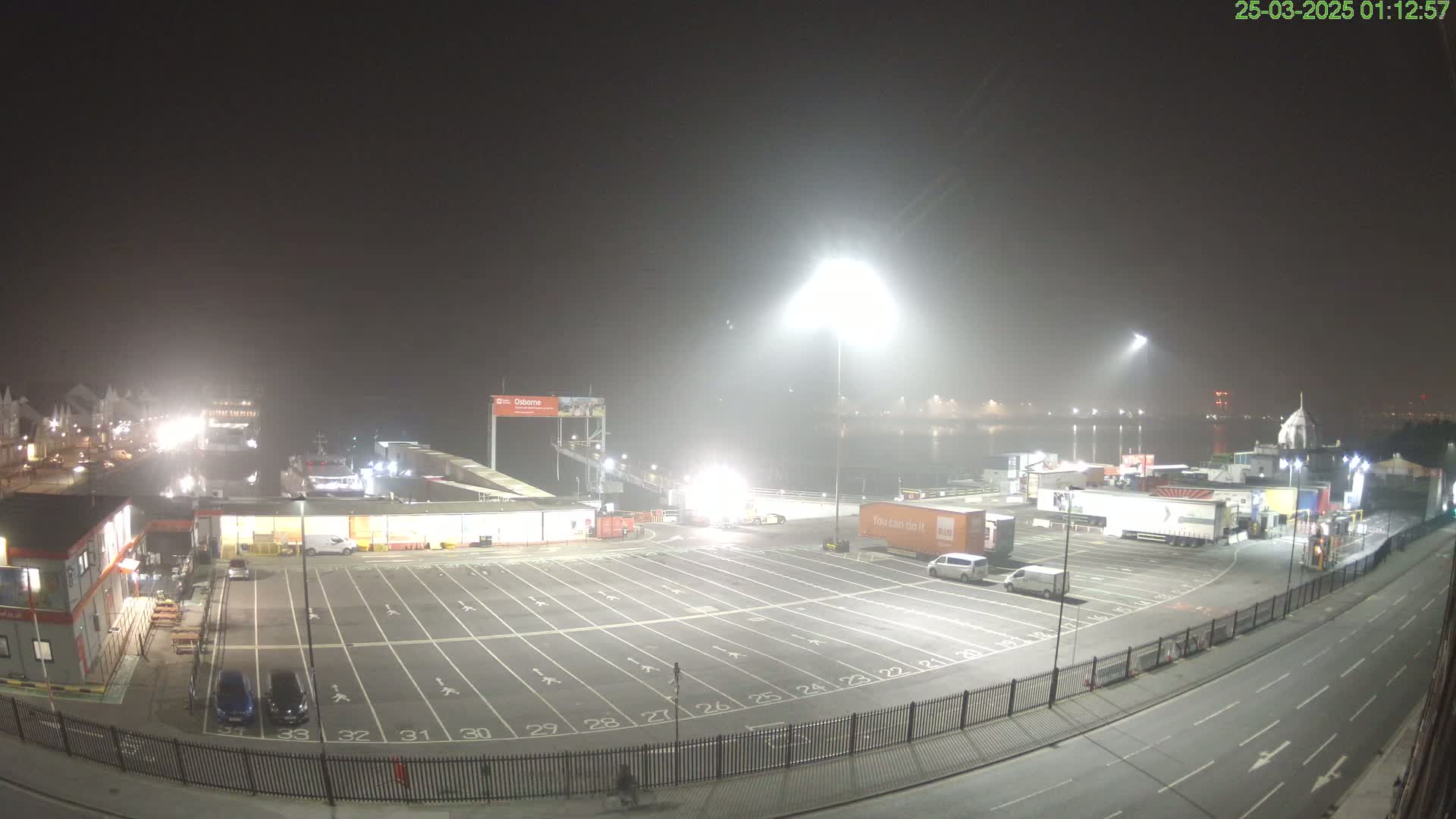A foggy nighttime view of a ferry terminal with an empty parking lot, several vehicles, and a ferry docked.