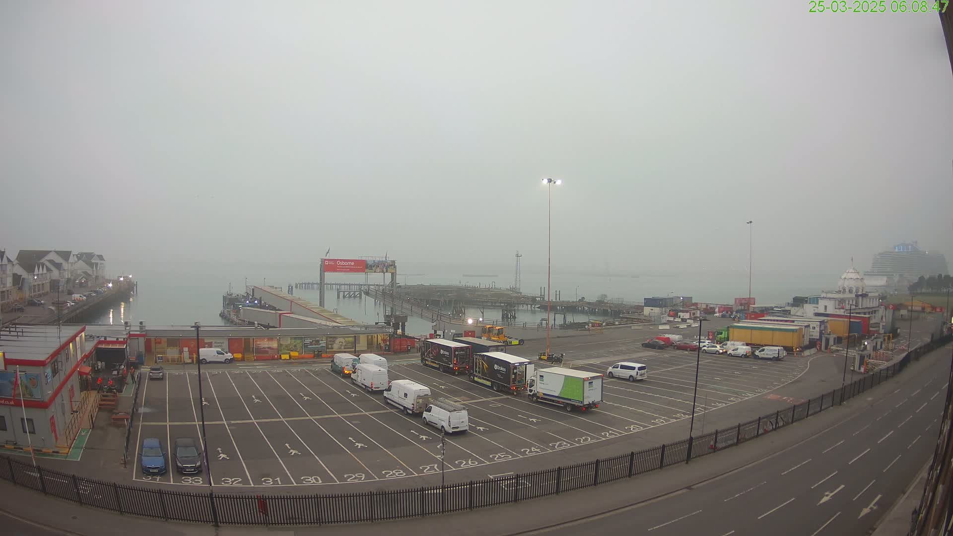 A foggy harbor scene shows a large, mostly empty parking lot filled with several delivery trucks and cars, next to a dock where a ferry is moored.