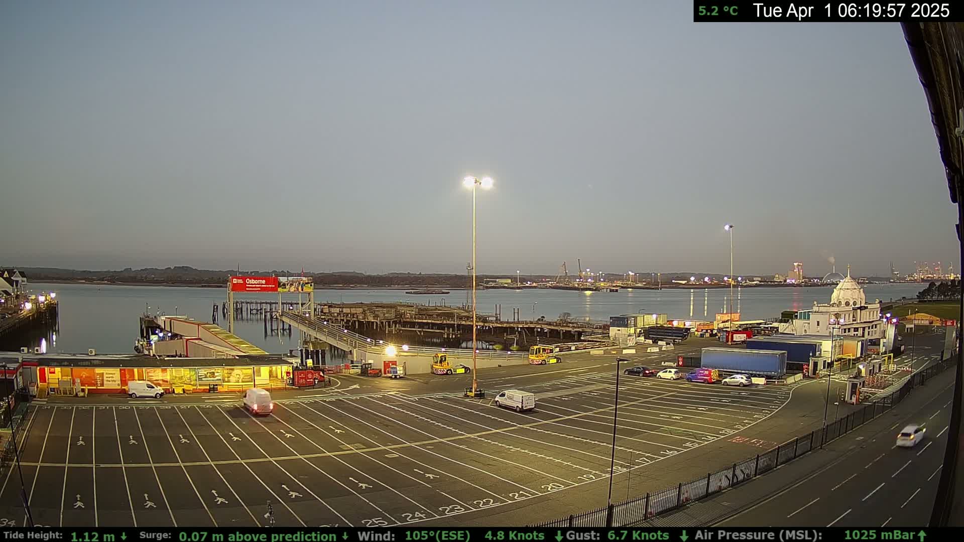 A mostly empty ferry terminal parking lot at dusk overlooks a calm body of water with distant city lights.