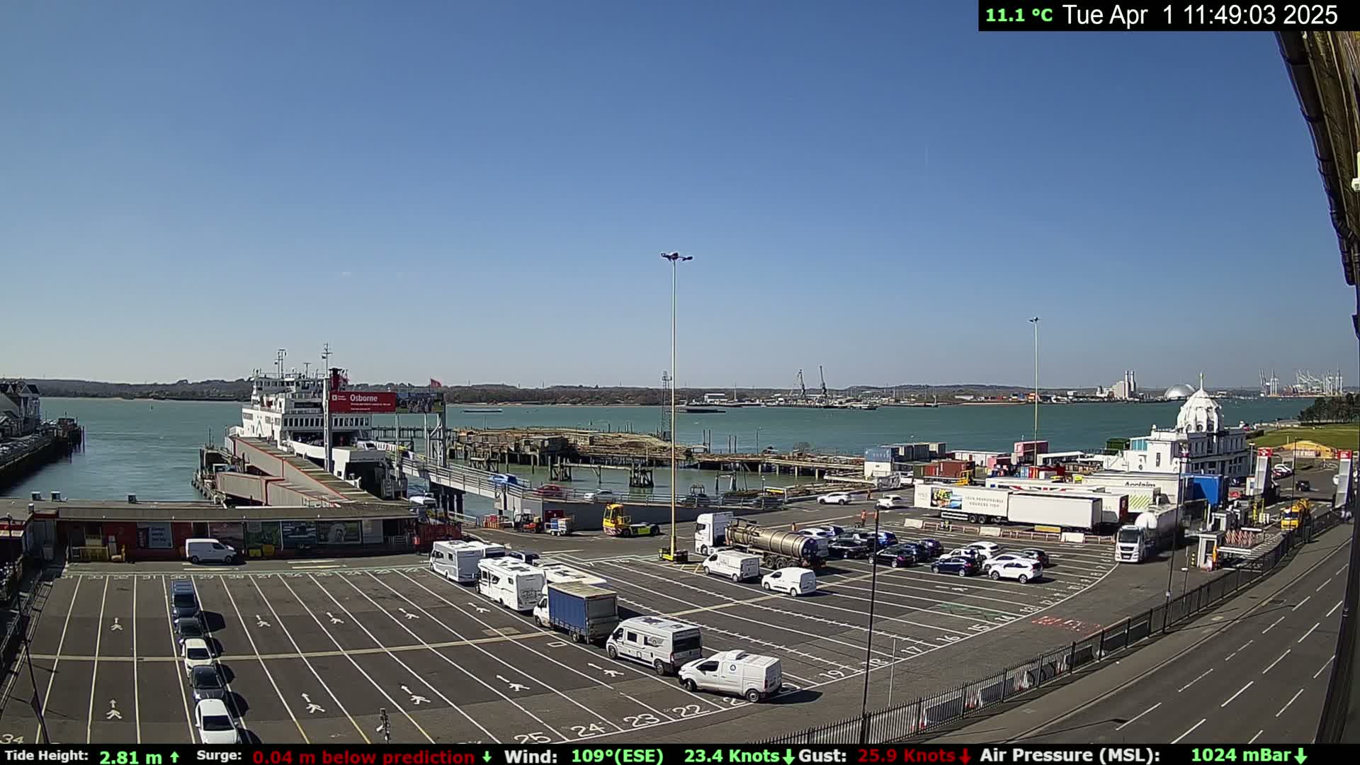 A ferry is docked at a terminal on a sunny day with a clear blue sky, and numerous vehicles are parked in the lot.