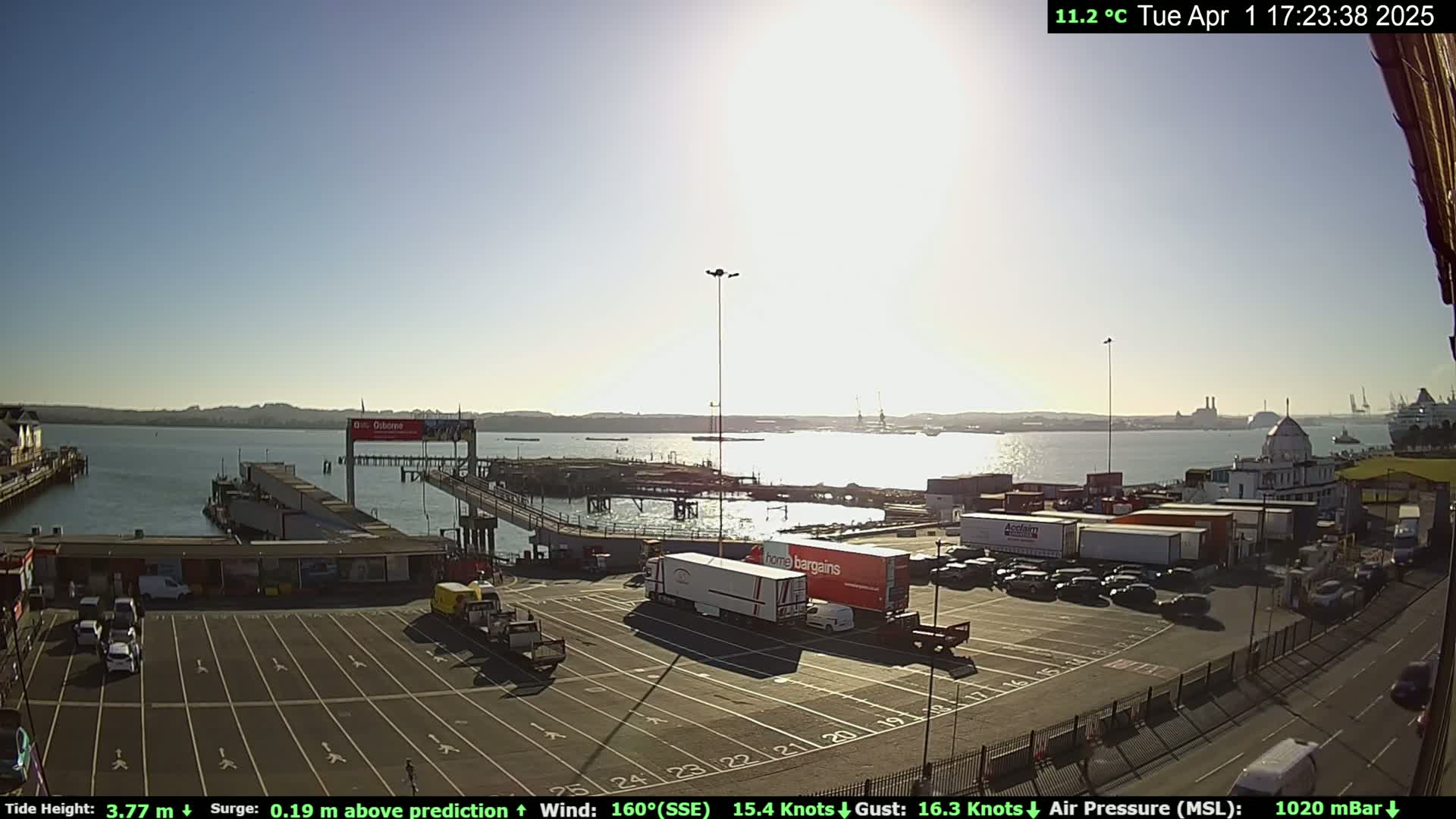 A sunny harbor scene shows a ferry terminal with parked cars and trucks, a calm body of water, and distant hills under a clear sky.