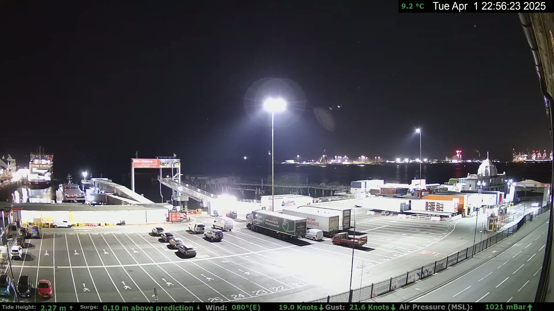 A nighttime view of a ferry terminal with several vehicles parked in the lot, a ferry docked, and other boats and structures visible across the water under a clear sky.