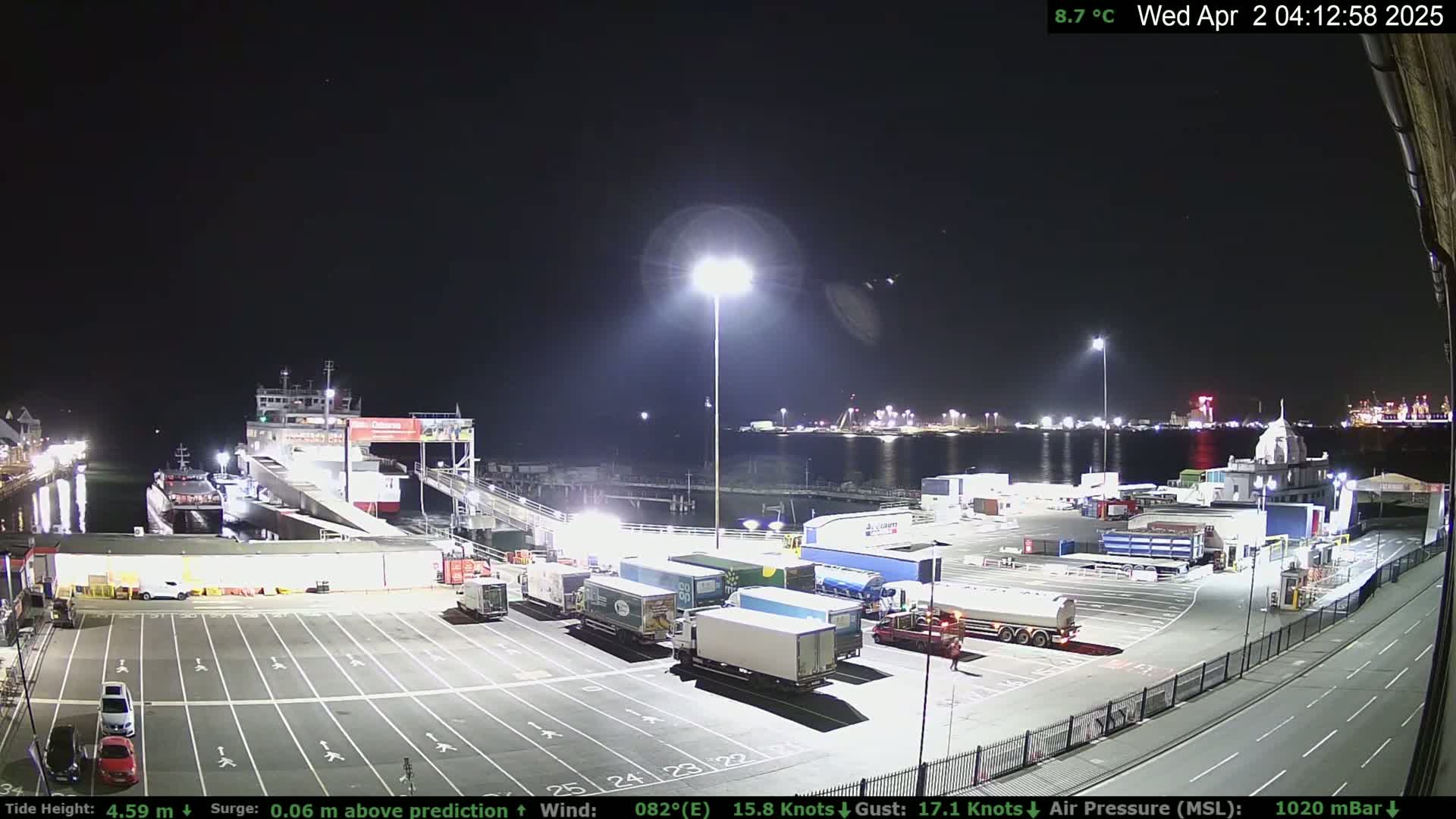 A nighttime view of a ferry terminal with several trucks parked in the lot, a ferry docked, and calm water in the harbor under a clear sky.