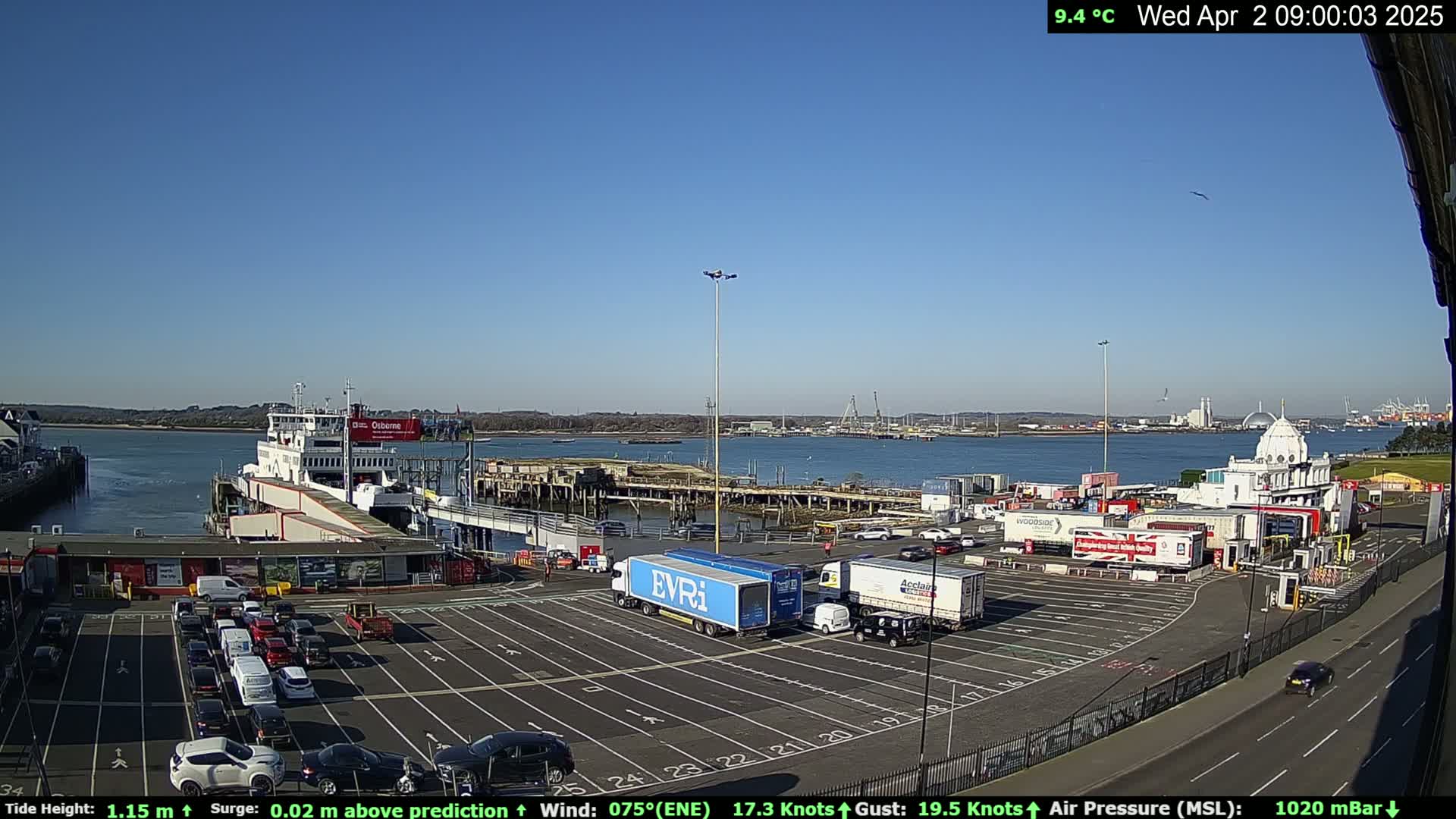 A ferry is docked at a terminal with numerous cars and trucks parked in a lot, under a clear blue sky.