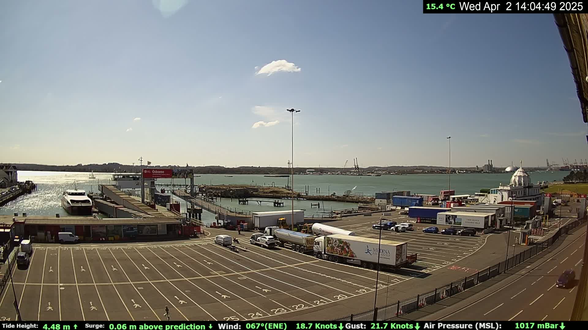 A ferry docks at a terminal on a sunny day with a light wind, while trucks and cars occupy a large parking lot nearby.